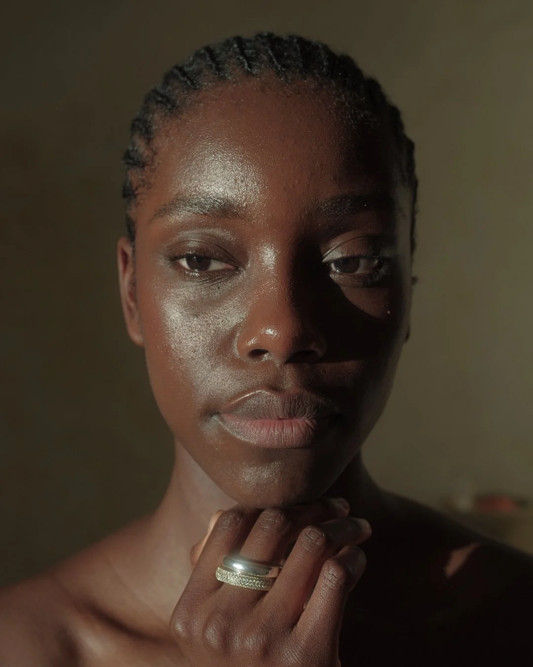 Close-up portrait of a young Black woman with natural hair, looking directly at the camera. She has smooth skin, subtle makeup, and is wearing a silver ring on her finger, which rests gently on her chin.