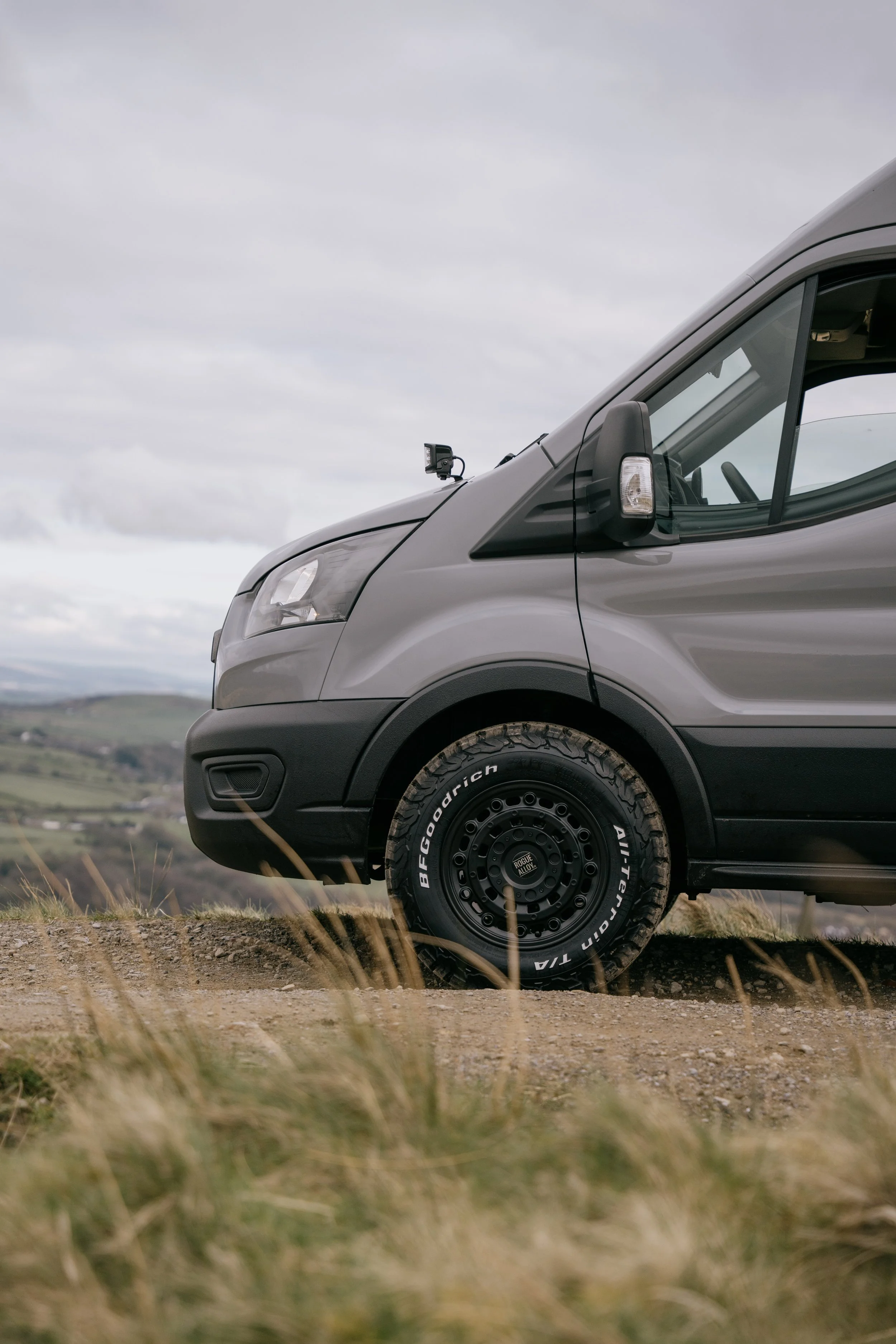 A grey van parked on a dirt road with hills and cloudy sky in the background.