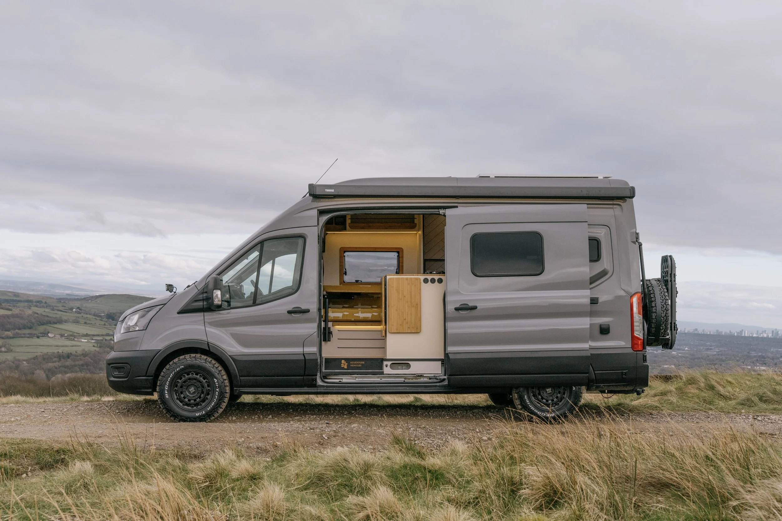 A grey Ford Transit camper van parked on a grassy hillside with an open side door revealing a small living space with a wooden interior, a window, and a kitchen area, against a cloudy sky and distant city skyline.