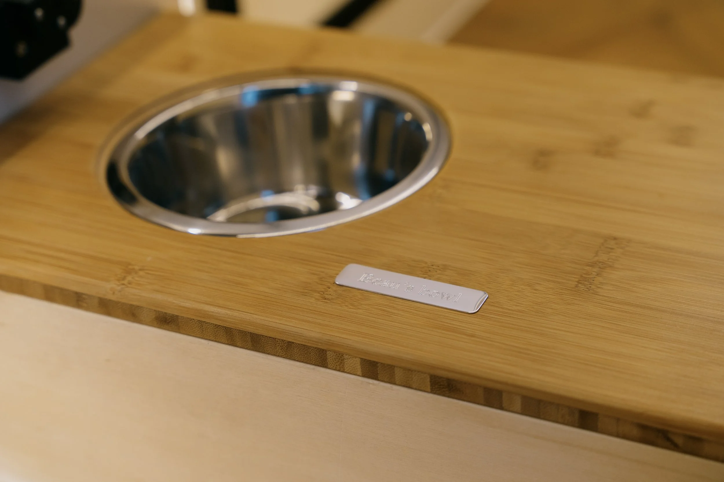 A small, round, stainless steel bowl embedded in a wooden surface, with a metal label that reads 'Feeding & Bowls' nearby.