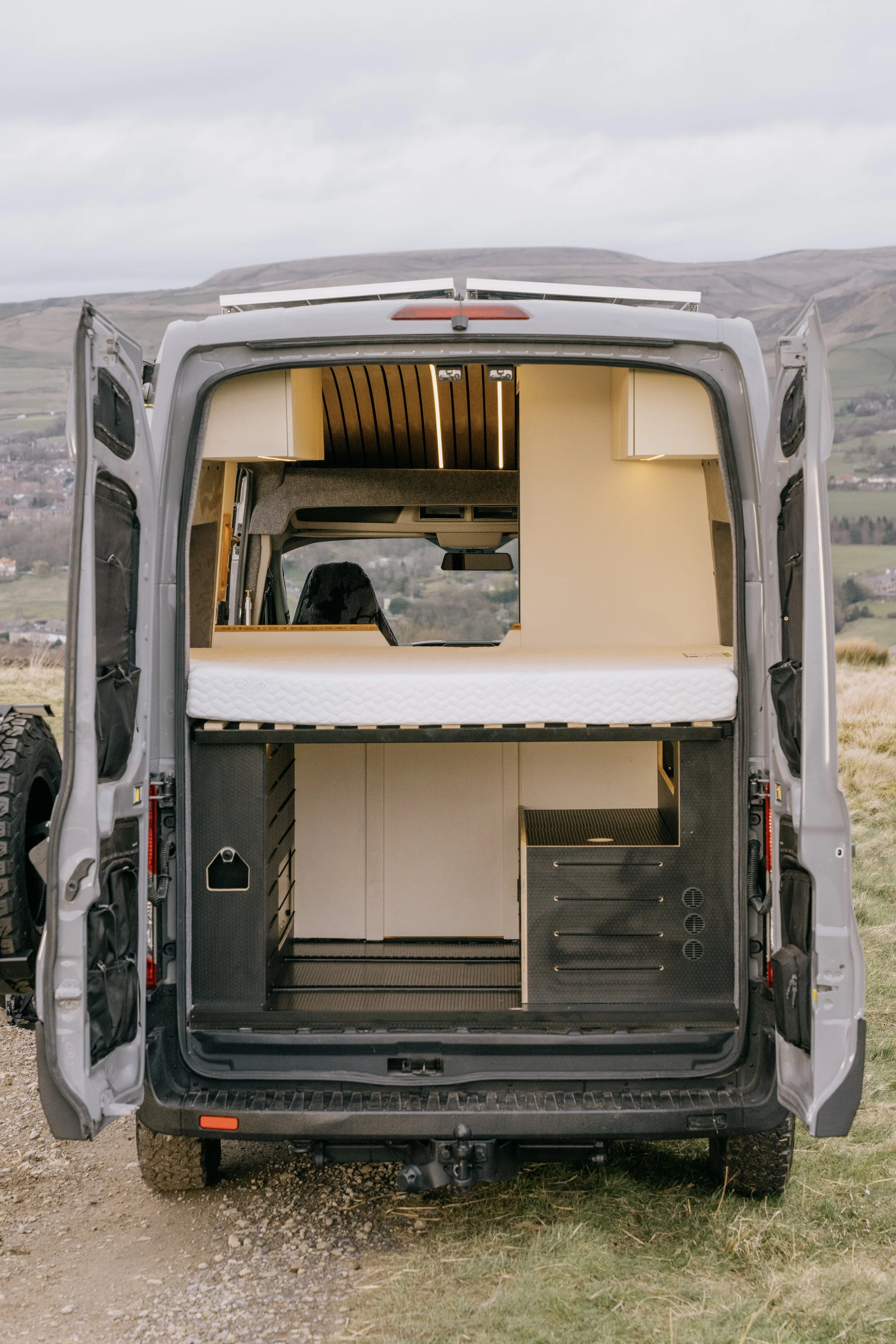 The back of a converted camper van with open doors, revealing an interior with storage compartments, a mattress, and wood paneling, situated outdoors on a grassy area with hills in the background.