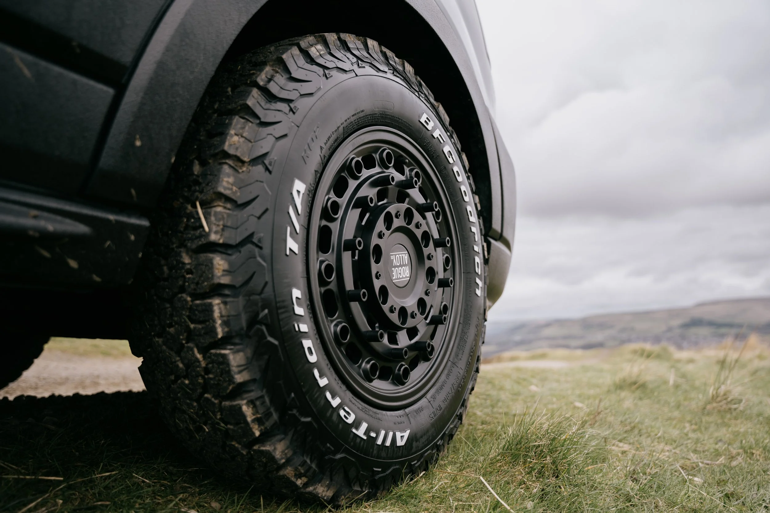 Close-up of a rugged off-road tire mounted on a black rim of a vehicle, on a grassy landscape with hills in the background, under a cloudy sky.
