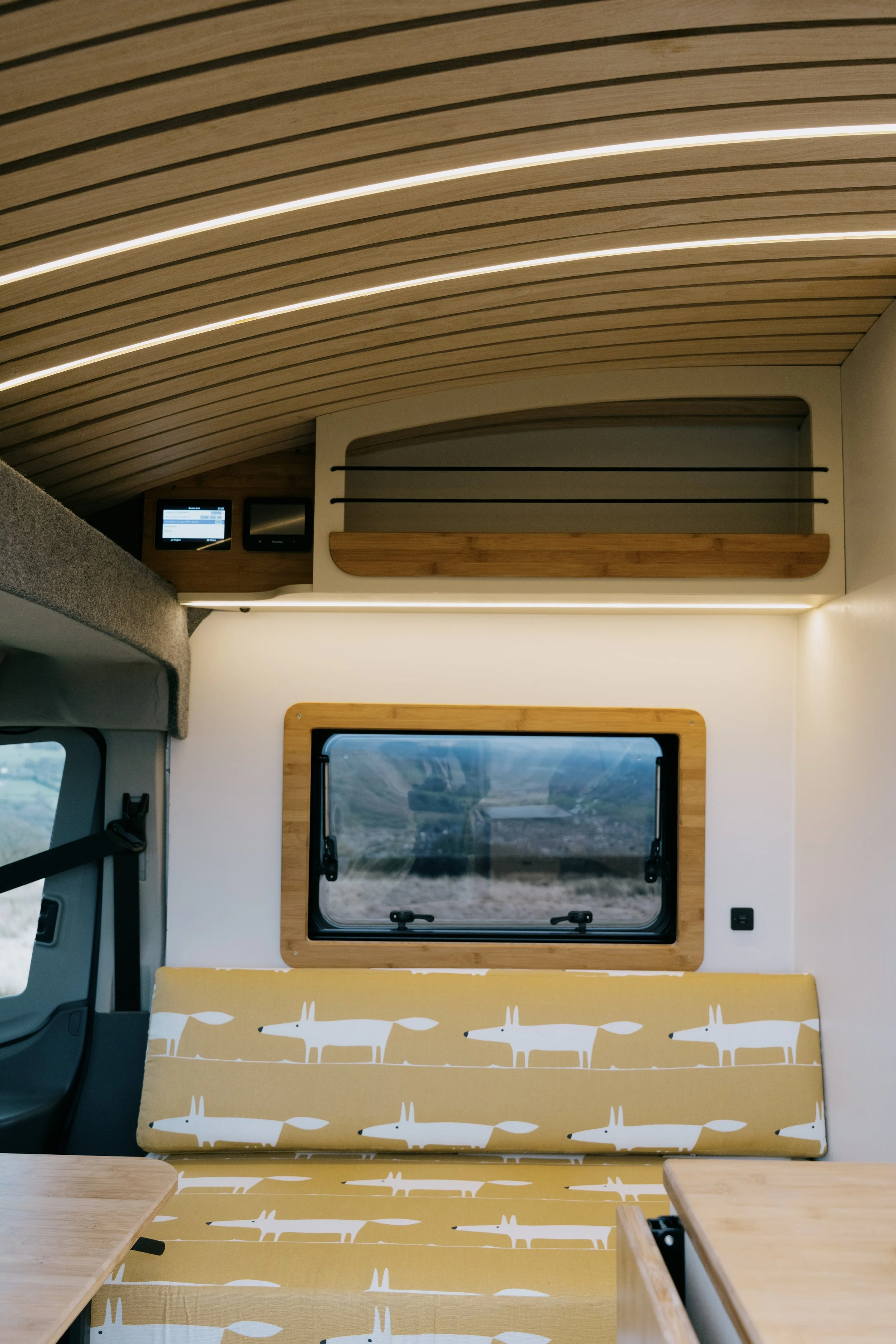 Interior of a camper van with a curved wooden ceiling, a window with a wooden frame, a patterned yellow and white seating area with foxes, and a small overhead storage compartment.