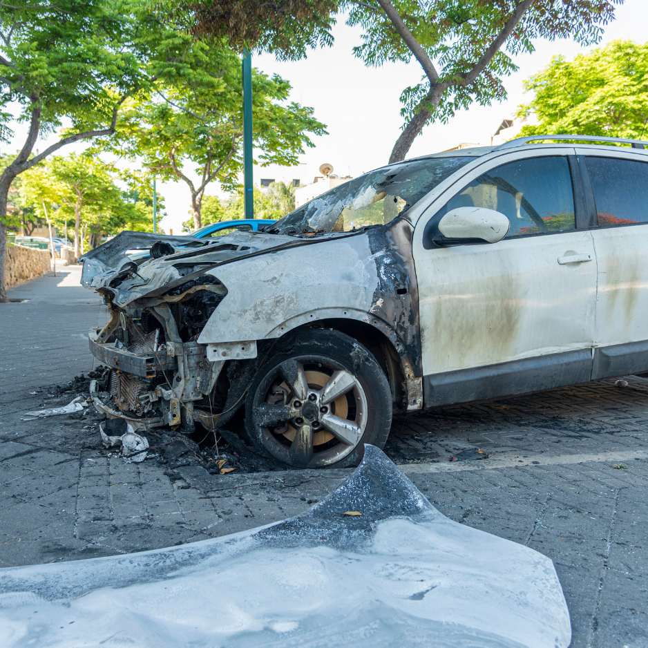 Burned-out car with severe front-end damage parked on a street, surrounded by trees.