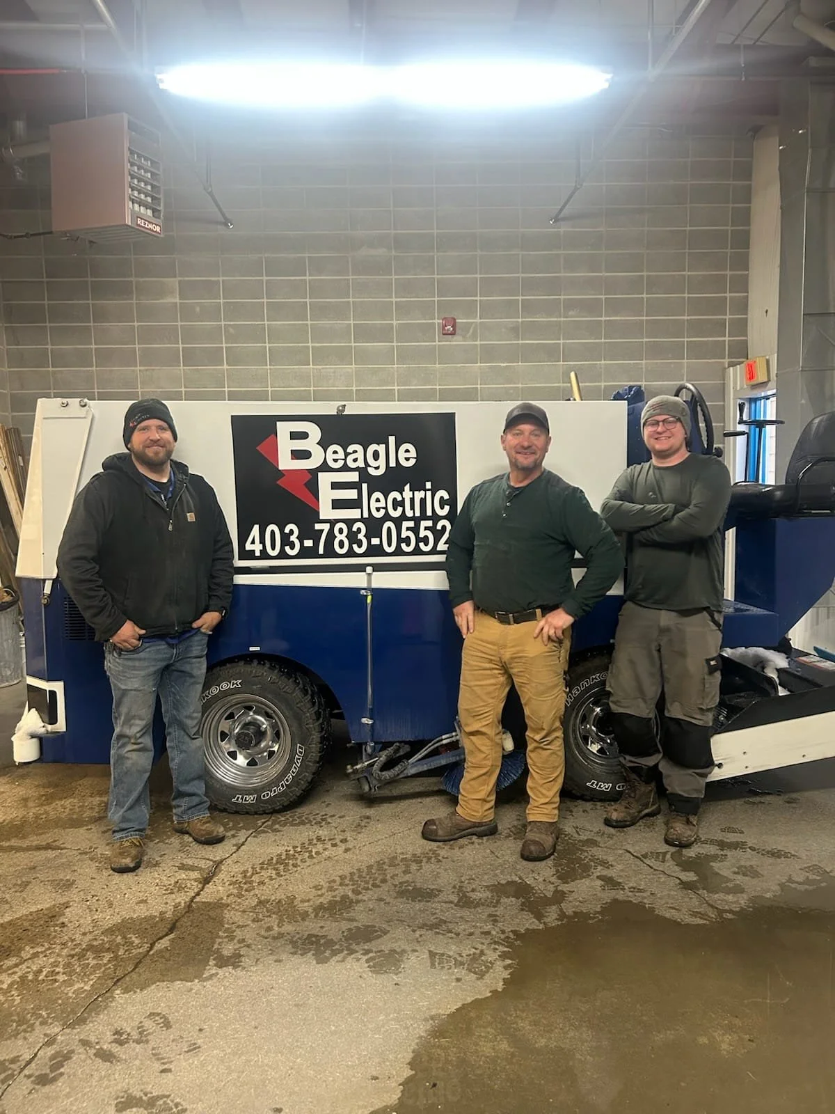 Three men standing in front of a blue utility vehicle with a sign that reads "Beagle Electric" and a phone number. They are inside a garage or workshop, wearing work clothes.