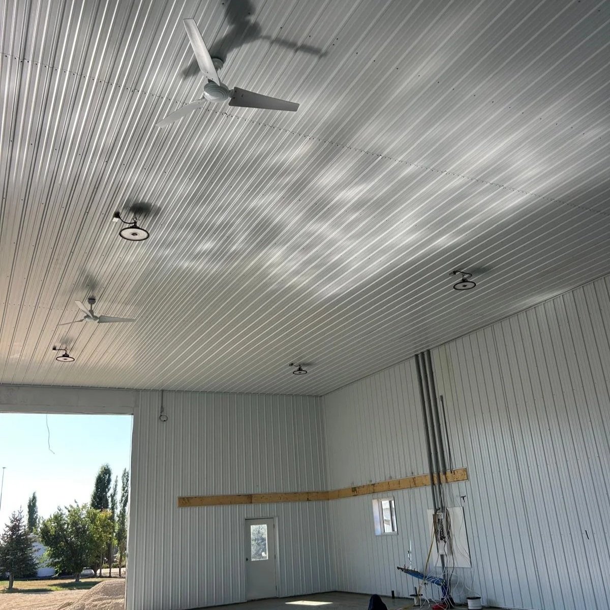 Interior view of a large metal building with a corrugated metal ceiling, ceiling fans, and industrial lighting fixtures. There is an open doorway showing trees outside.