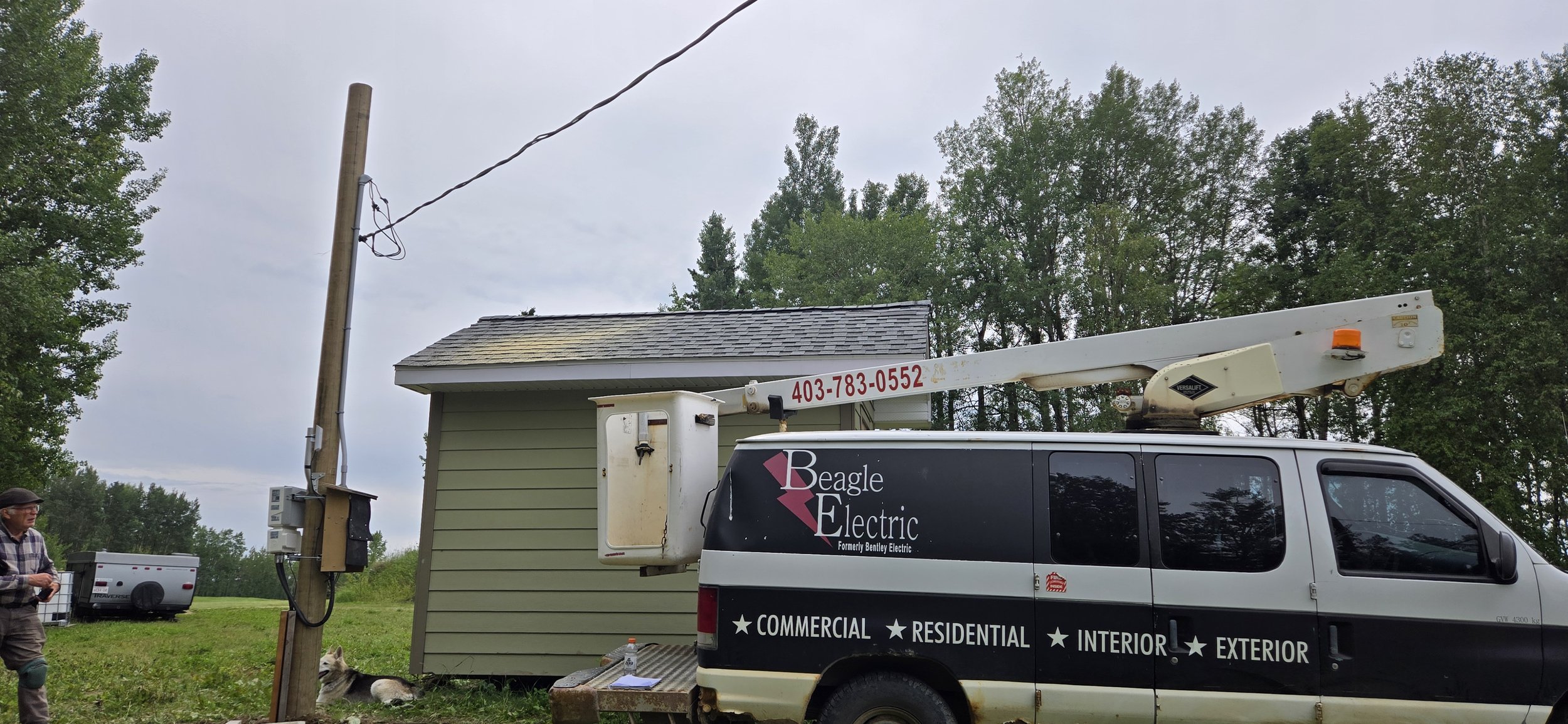 Electrician's van labeled 'Beagle Electric' with the phone number 403-783-0552, parked next to a house with a cherry picker lift. There is a man and a dog in the grassy yard and trees in the background.