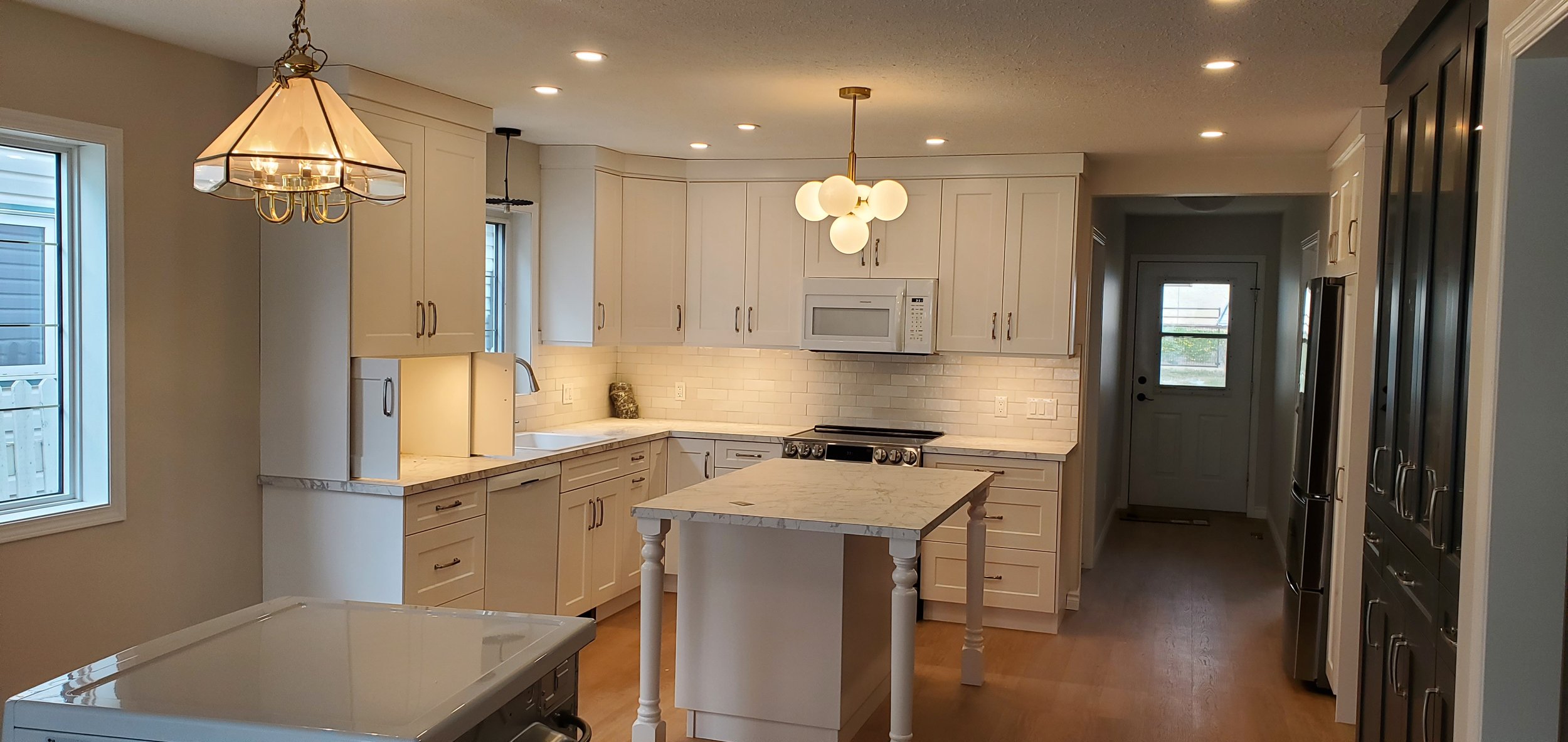 Modern kitchen with white cabinets, marble countertops, and a small central island. There are two pendant lights hanging from the ceiling, and a row of recessed lights. A microwave is built into the cabinetry above the stove, and there are windows on the left and a door at the back of the kitchen.