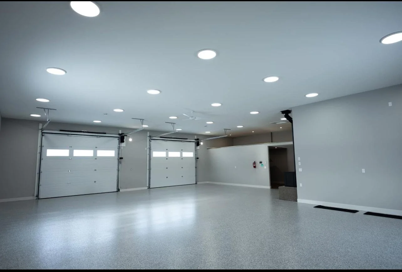 Empty residential garage with two closed garage doors, bright ceiling lights, light gray walls, and a speckled epoxy floor.