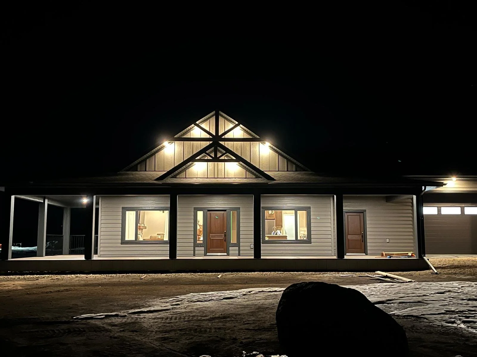 A house illuminated at night, featuring a front porch, central wooden door, two windows, and a garage on the right, with construction materials visible outside.