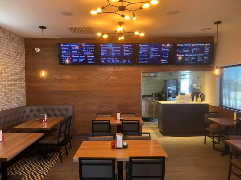 Interior of a modern restaurant with wooden tables and chairs, a brick wall on the left, digital menu screens on the back wall, and a kitchen visible through a pass-through window.
