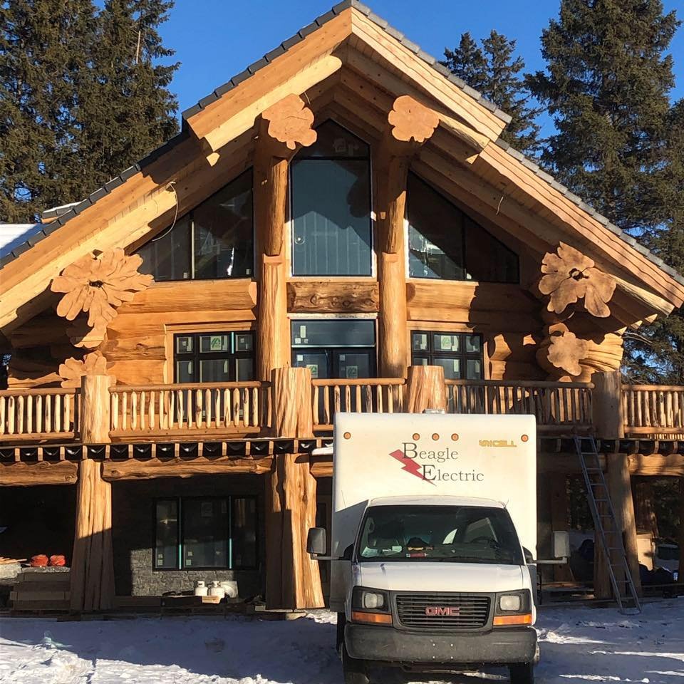 A two-story wooden house under construction with large log beams and decorative wooden flower accents on the facade. A white utility truck labeled "Beagle Electric" is parked in front, on a snowy ground, with a ladder leaning against the house.