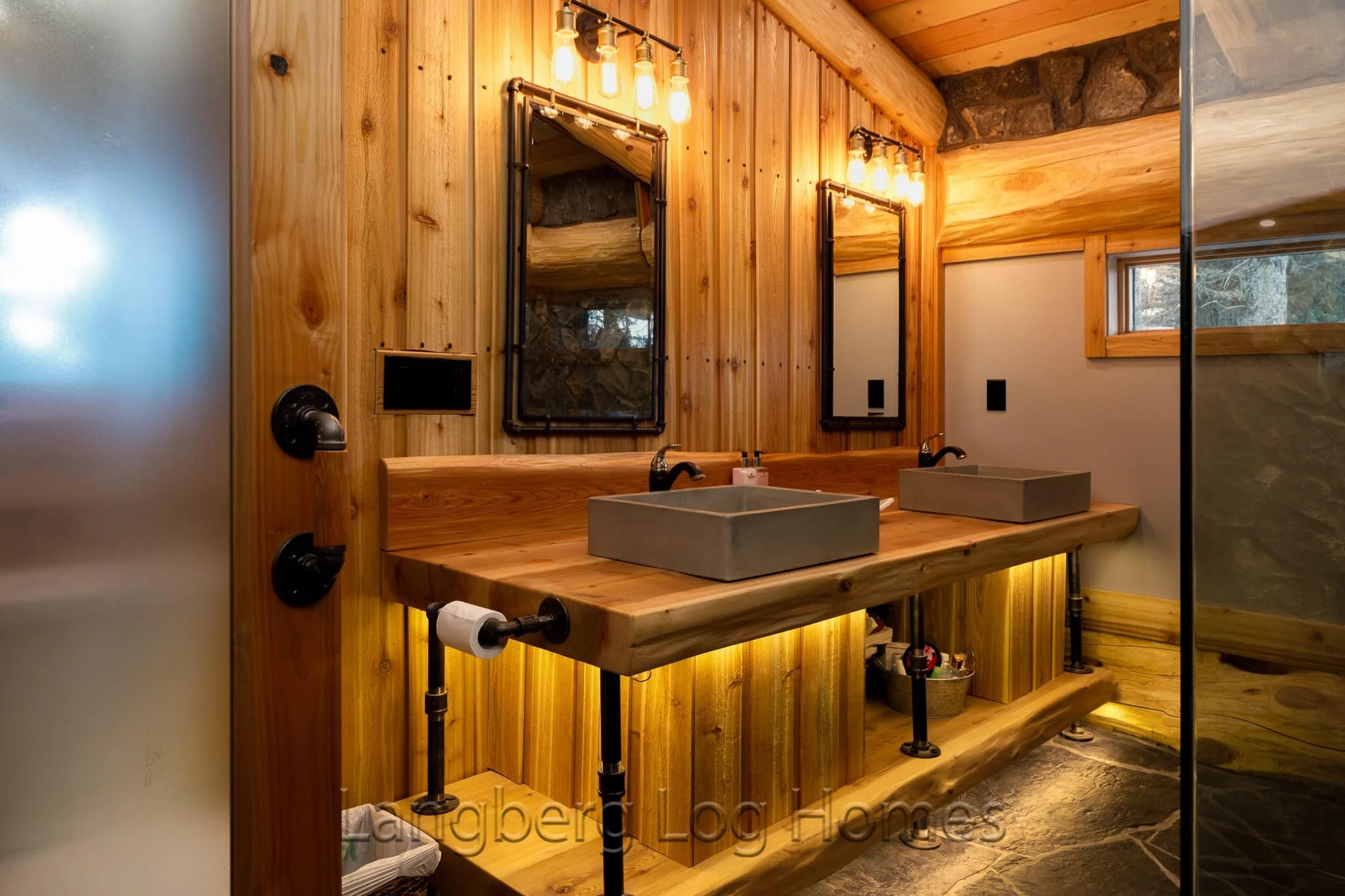Rustic bathroom with dual sinks, wooden vanity, and industrial-style mirrors with warm Edison bulb lighting.