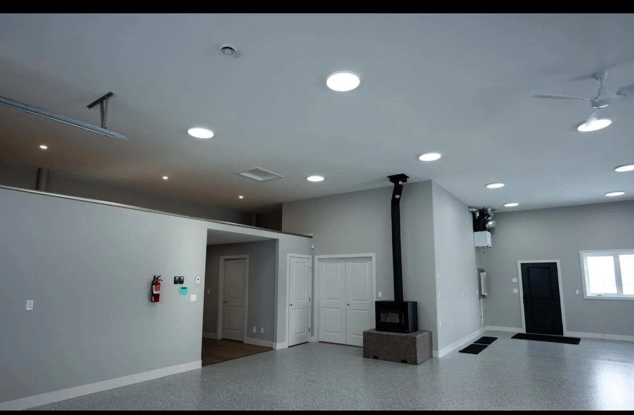 Interior of a modern, minimally furnished living space with gray walls, ceiling lights, a black door, a window, a ceiling fan, and a fireplace with a chimney.