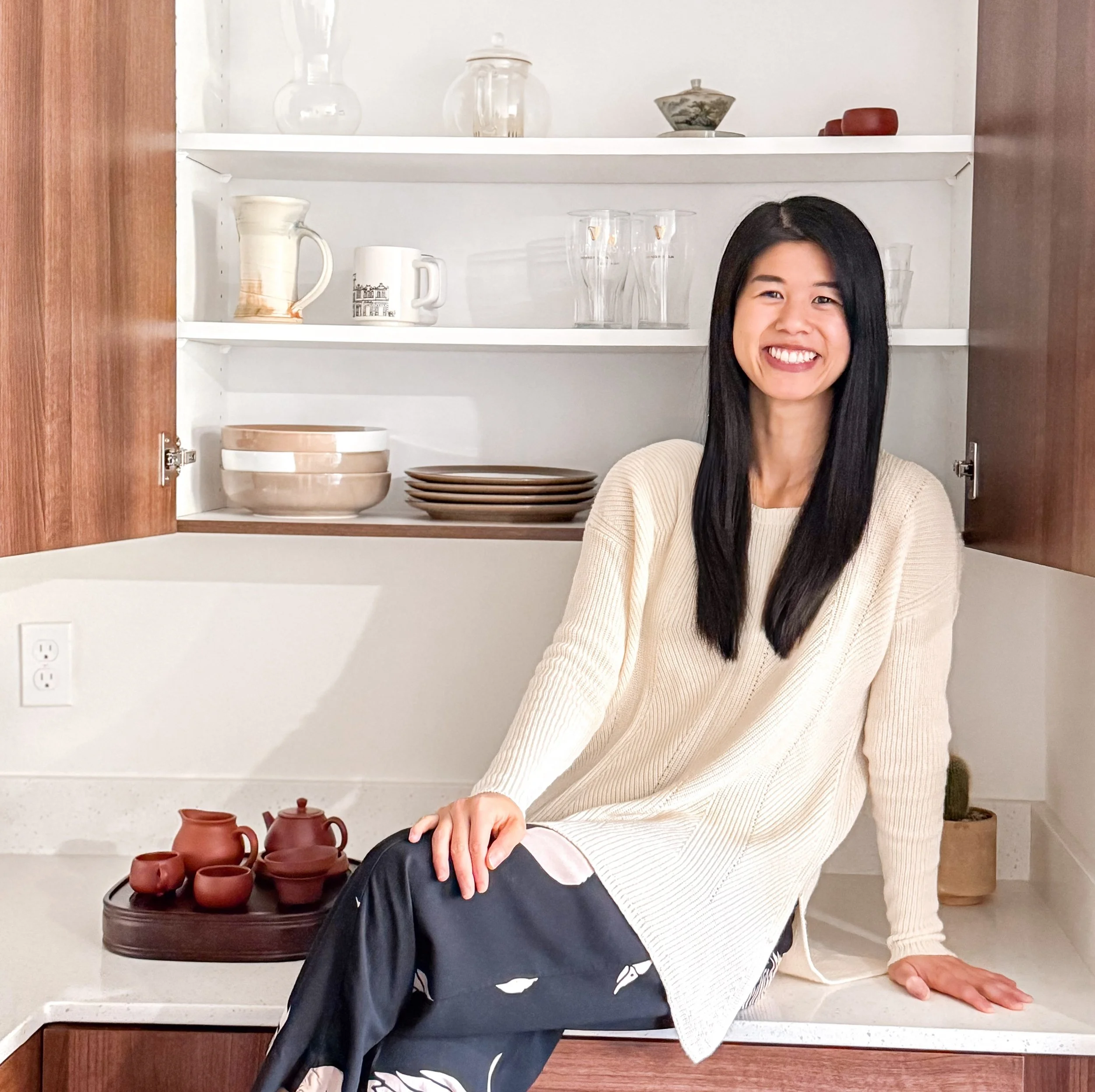 Owner, Noel sitting on a kitchen countertop in front of open cabinets filled with dishes and glassware organized on the shelves. Small potted plant and full tea set displayed on the counter.