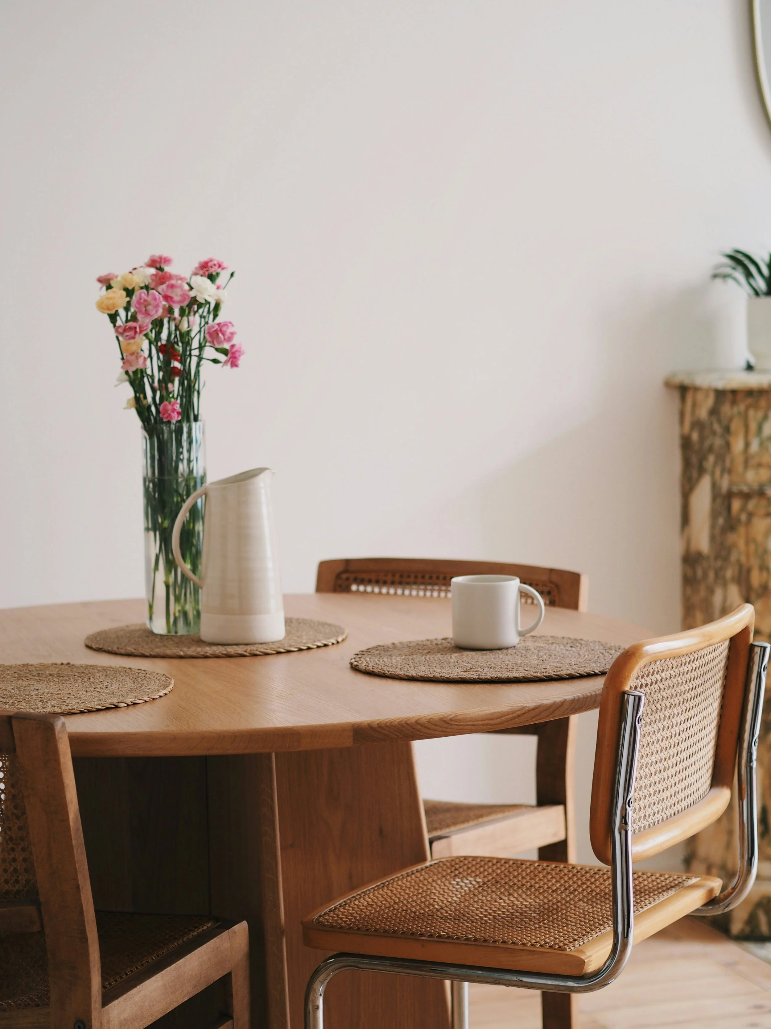 A minimalist dining room featuring a clear round wooden table, three woven chairs, and a small vase of pink flowers. This space illustrates the KonMari Method in Chehalis, Washington, showing how clear surfaces can calm the nervous system