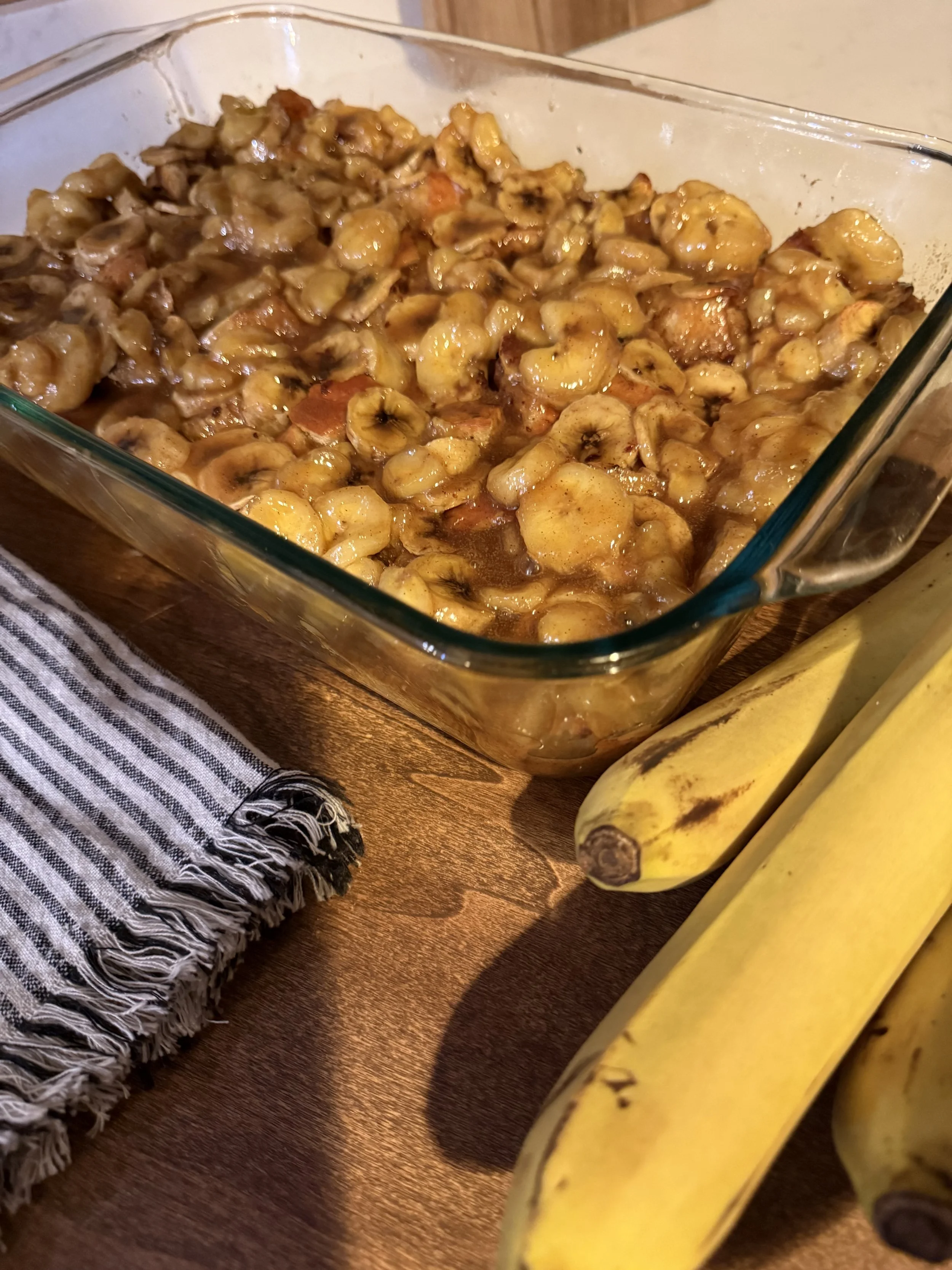 The most rich, buttery, and caramel-like bread pudding you'll ever taste, sitting on a wooden tray with bananas all around.
