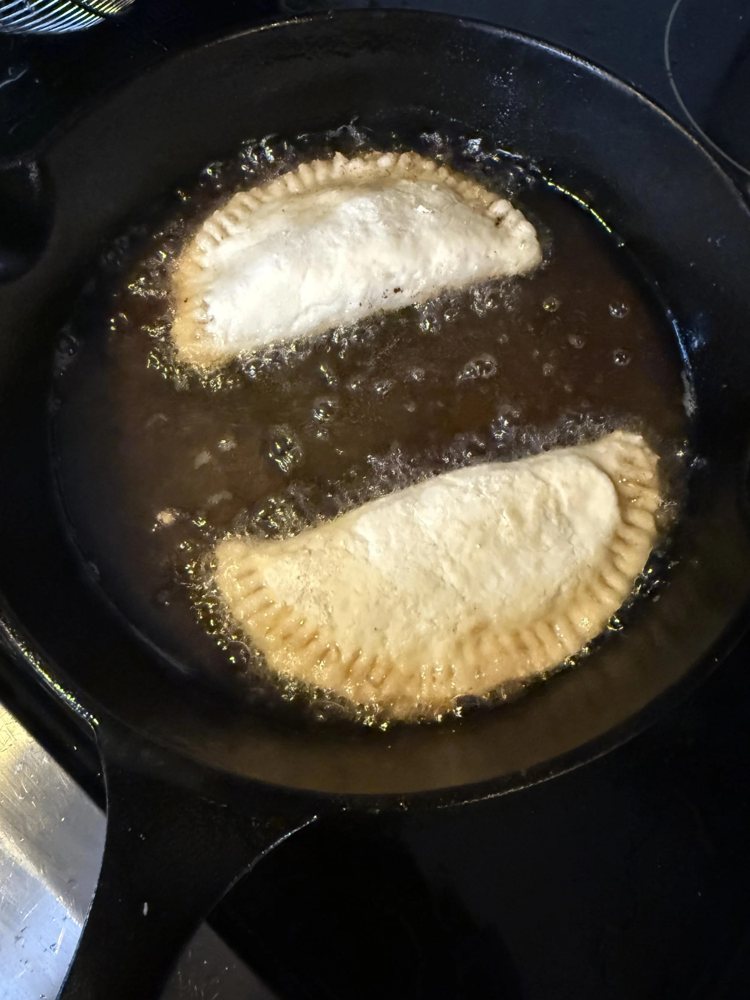 Two crimped apple pies frying in bubbling oil in a cast-iron skillet, with their edges turning golden.