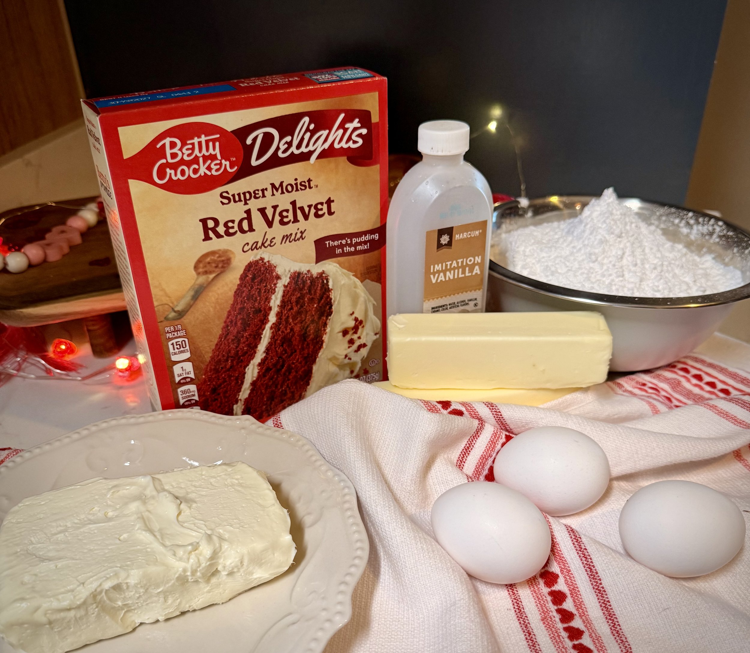 Ingredients for red velvet gooey butter cake bars including red velvet cake mix, cream cheese, eggs, butter, vanilla extract, and powdered sugar arranged on a farmhouse kitchen counter.