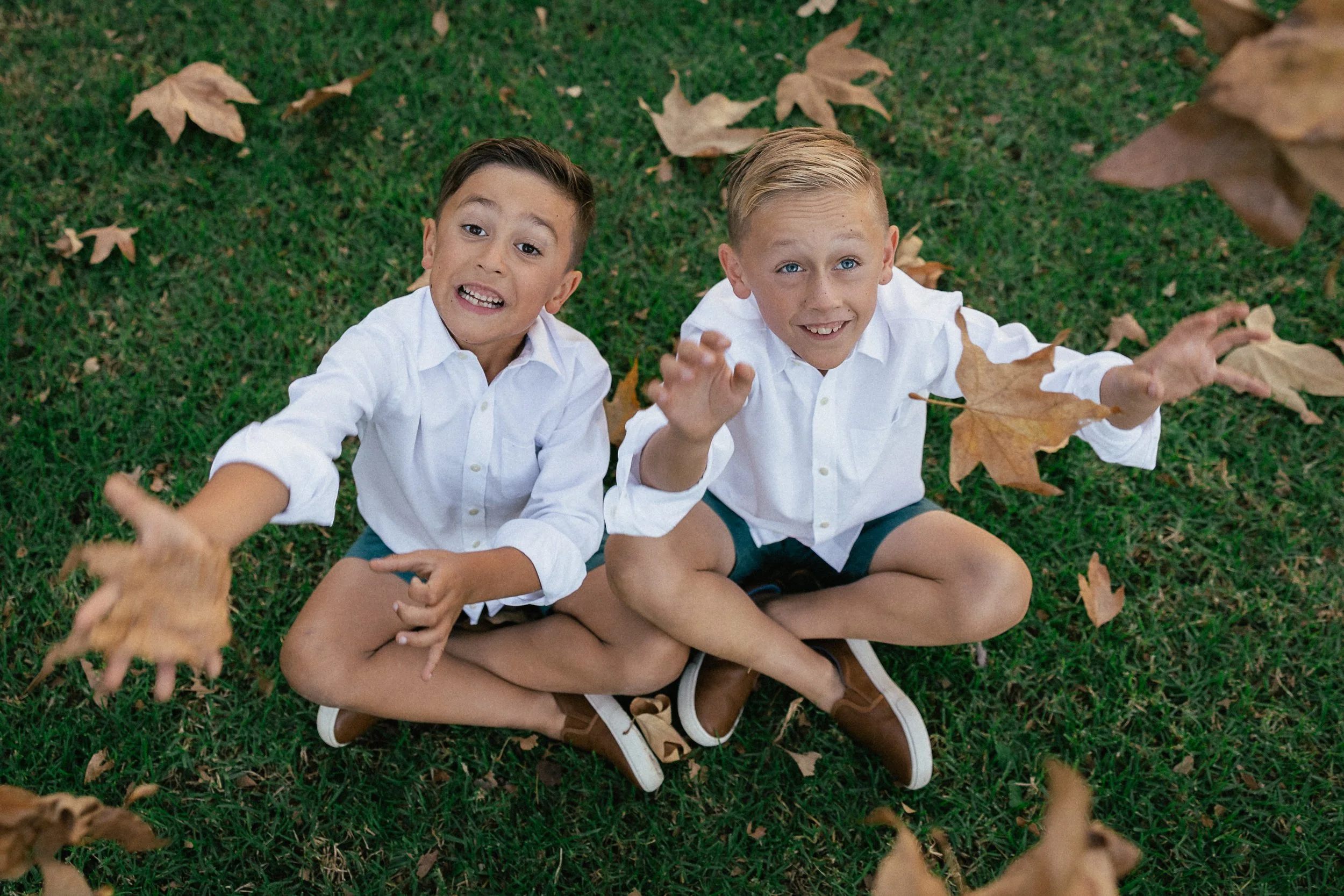 Two boys sitting on the grass and tossing leaves up toward the camera.