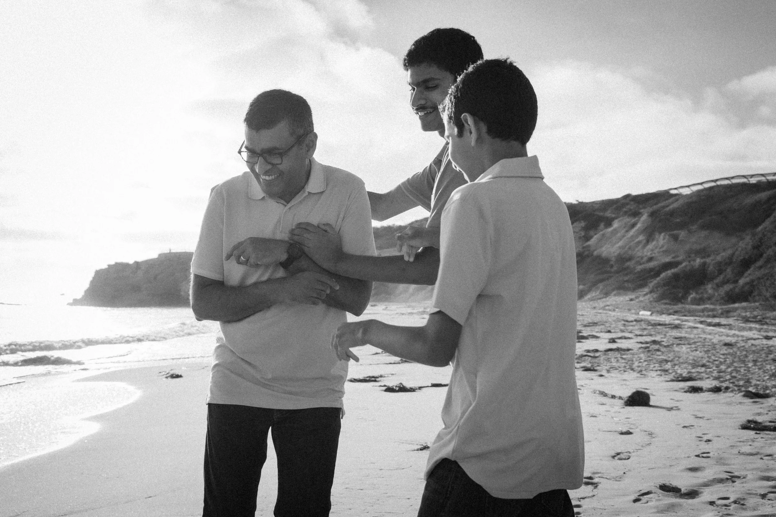 Candid black and white photo of father and two sons laughing on the beach at Crystal Cove in Orange County.