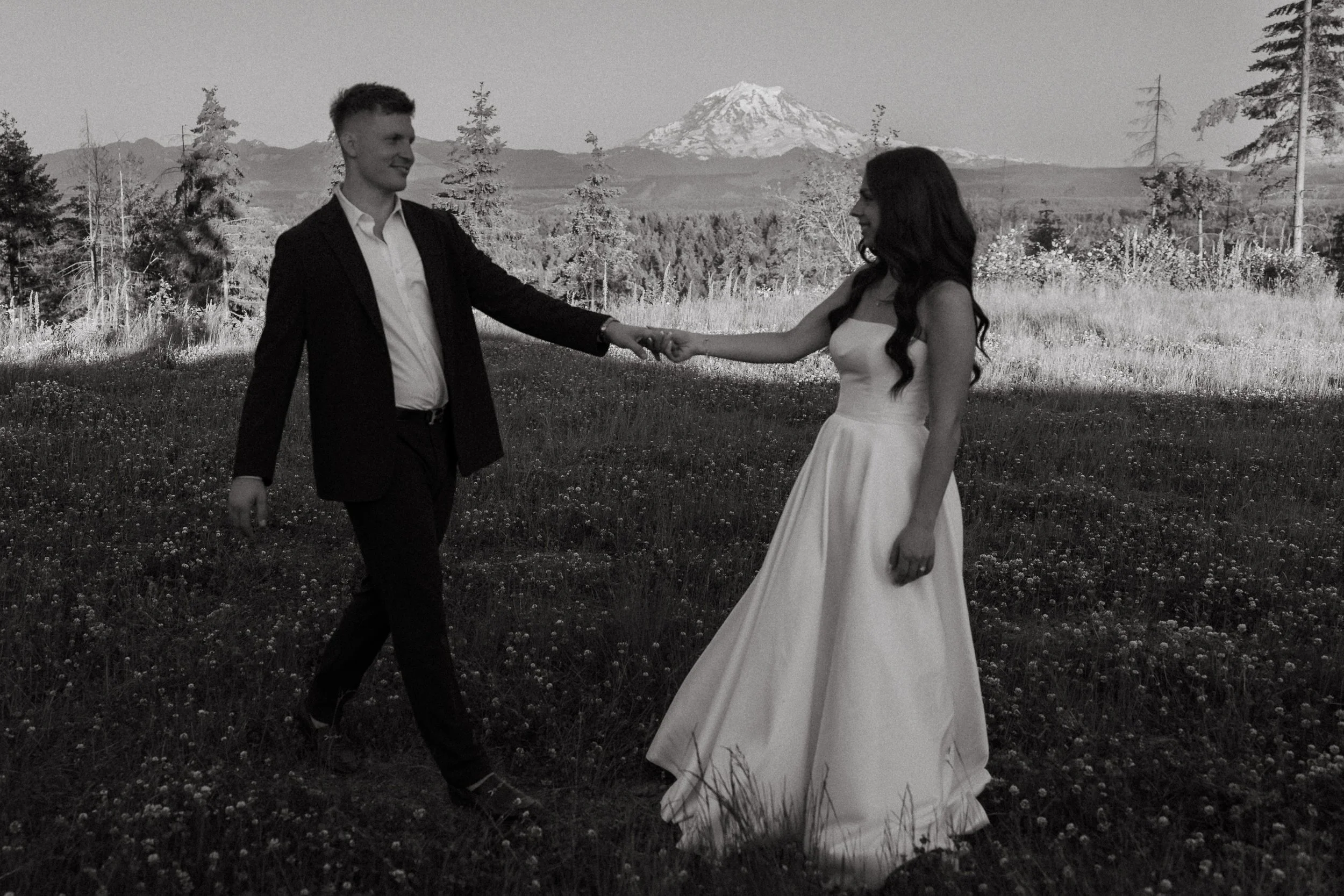 Bride and groom walking hand in hand through a mountain field with Mount Rainier in the distance.