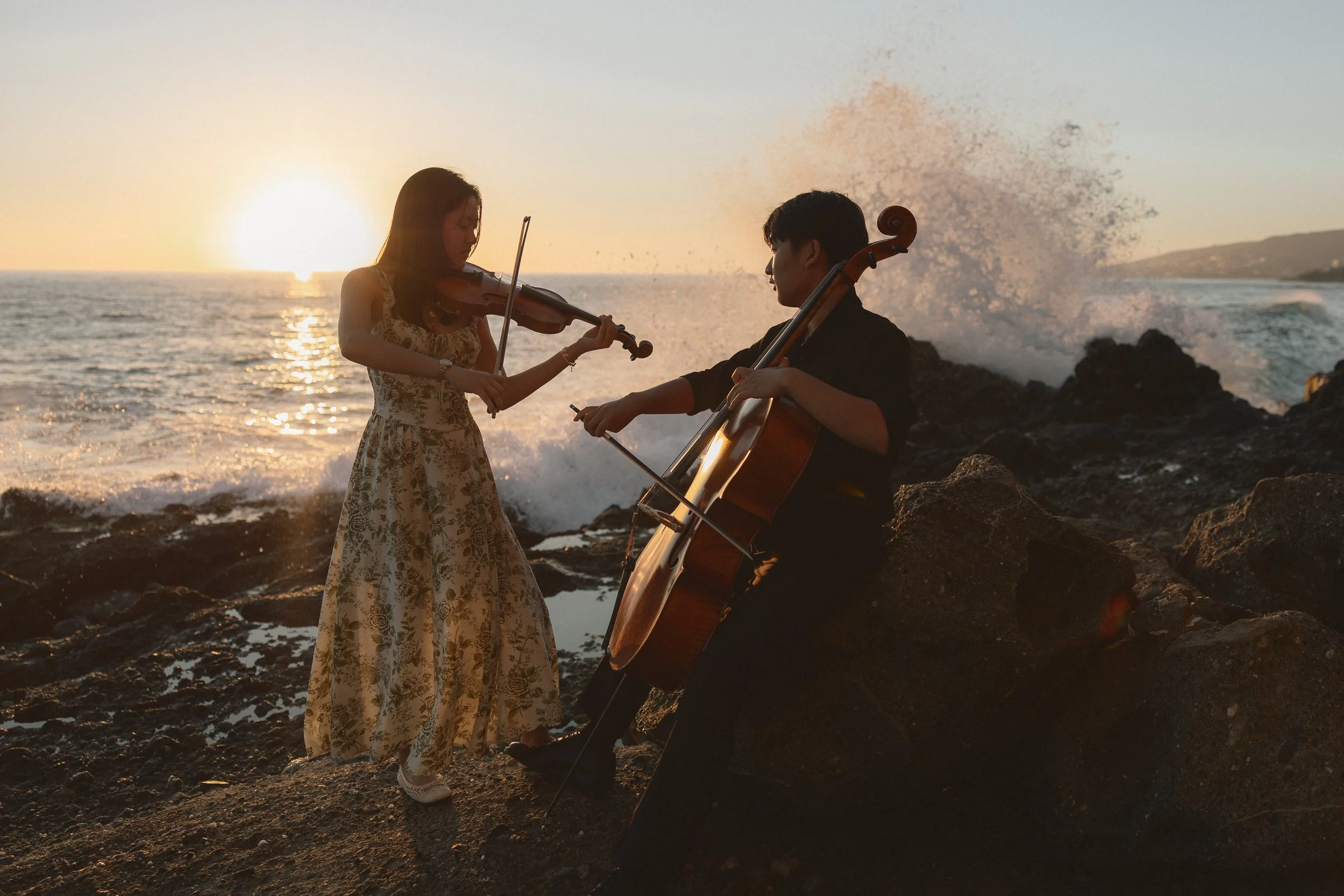A dramatic senior portrait of two musicians playing cello and violin on the ocean rocks at Treasure Island Beach during sunset.