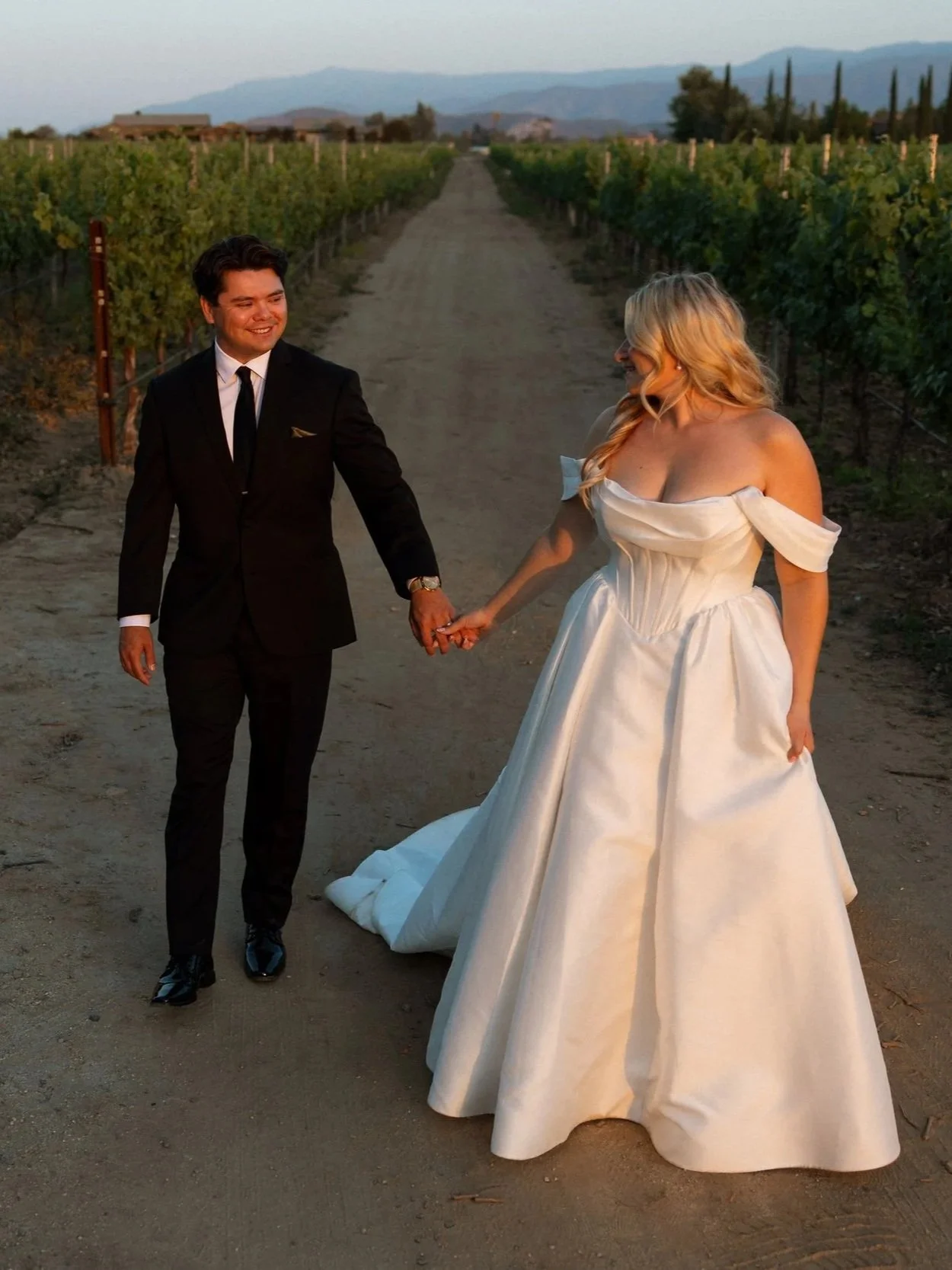Bride and groom hold hands walking down a vineyard row at sunset