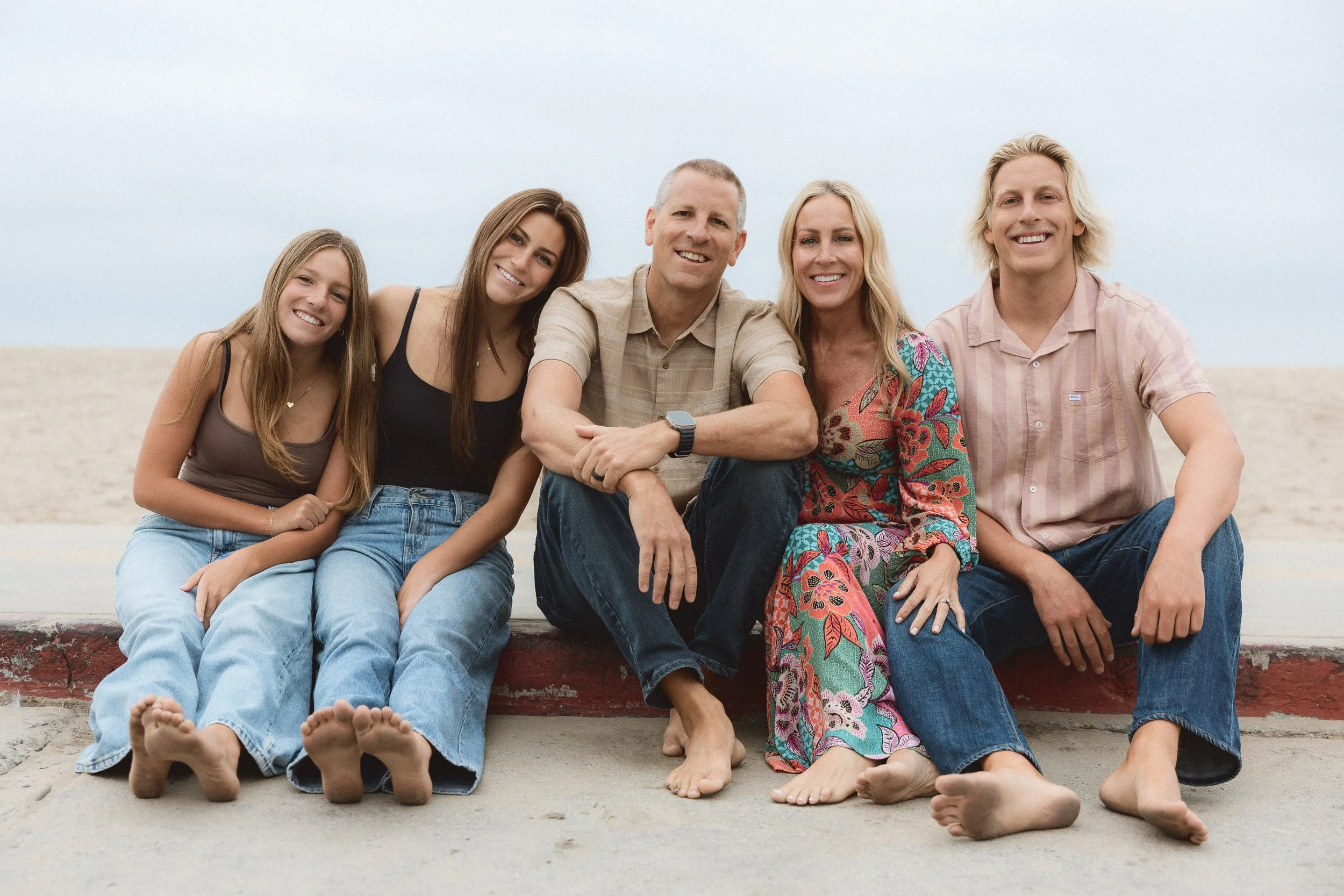 A family of five sitting barefoot on a concrete curb near the sand during a relaxed beach session on the Balboa Peninsula in Newport Beach.