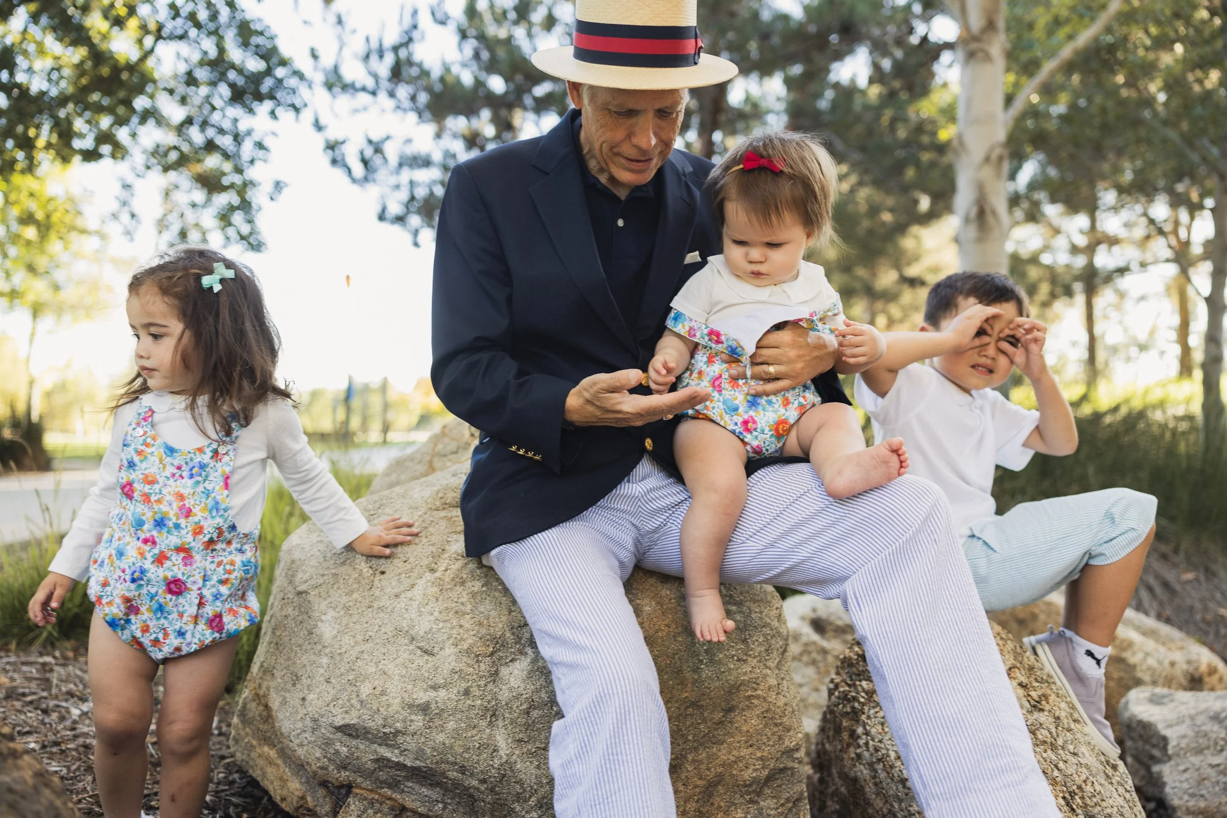 A grandfather sitting on a rock holding a baby while grandchildren play nearby during an extended family session at Jeffrey Open Space Trail in Irvine.