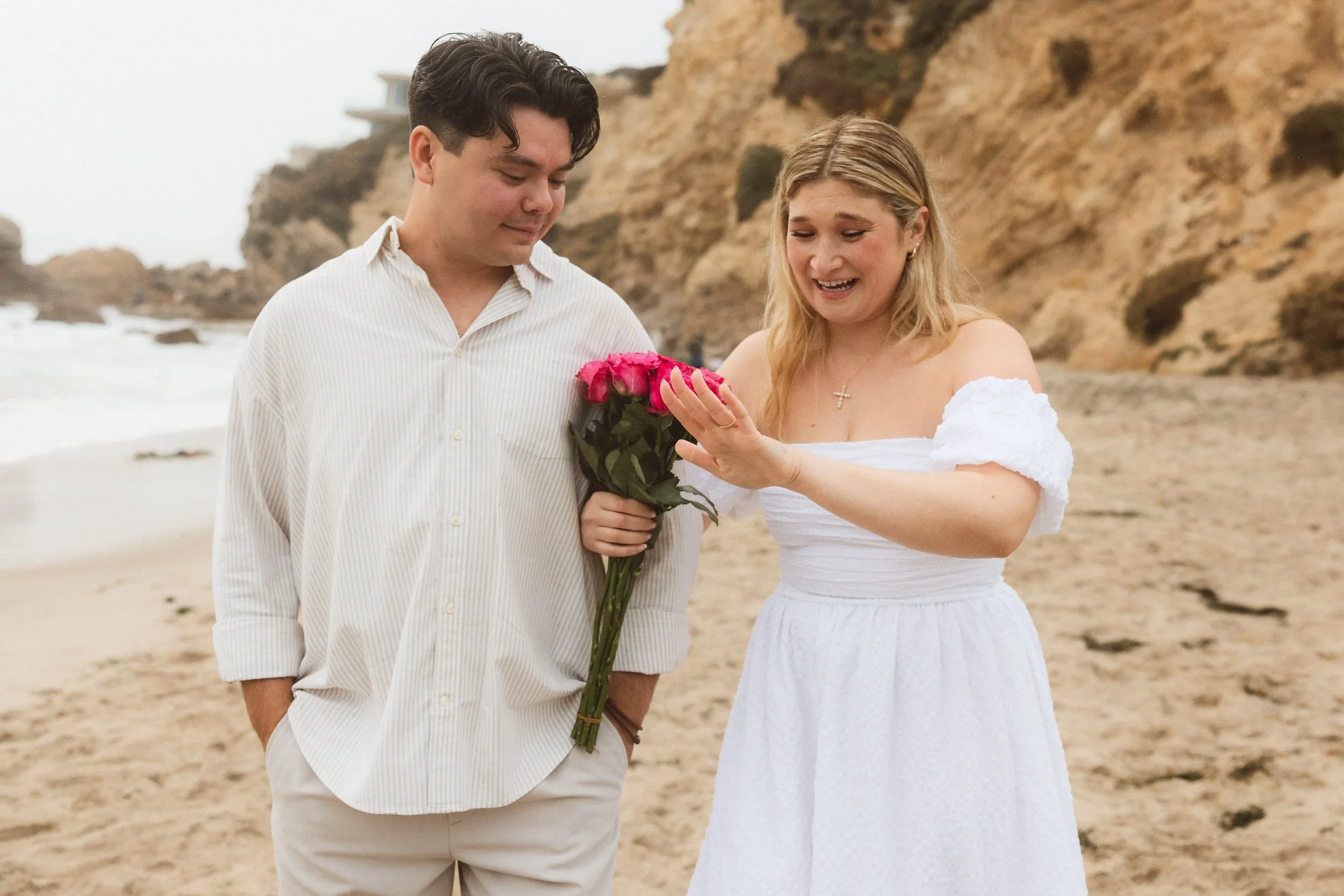 A joyful woman reacting with surprise while holding a bouquet of red roses after a beach proposal at Little Corona Del Mar in Newport Beach.