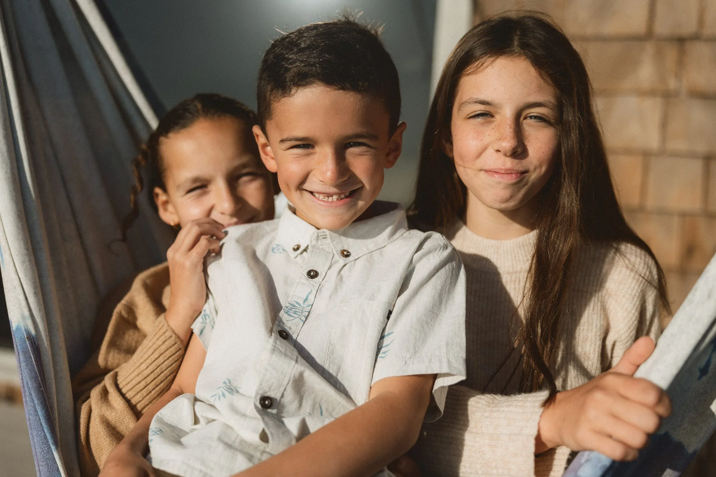 Two siblings laughing and hiding behind a hammock on a wooden deck during a relaxed family session at Capistrano Beach.