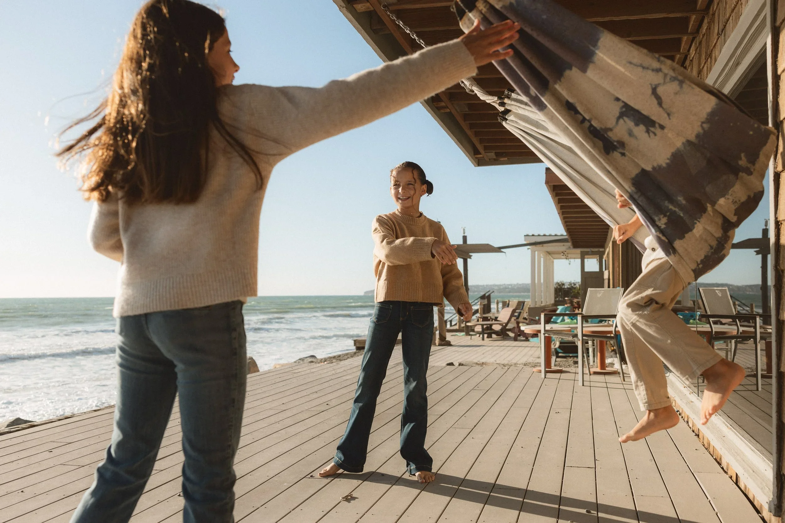 Two siblings laughing and hiding behind a patterned beach blanket on a wooden deck during a candid family session at Capistrano Beach.