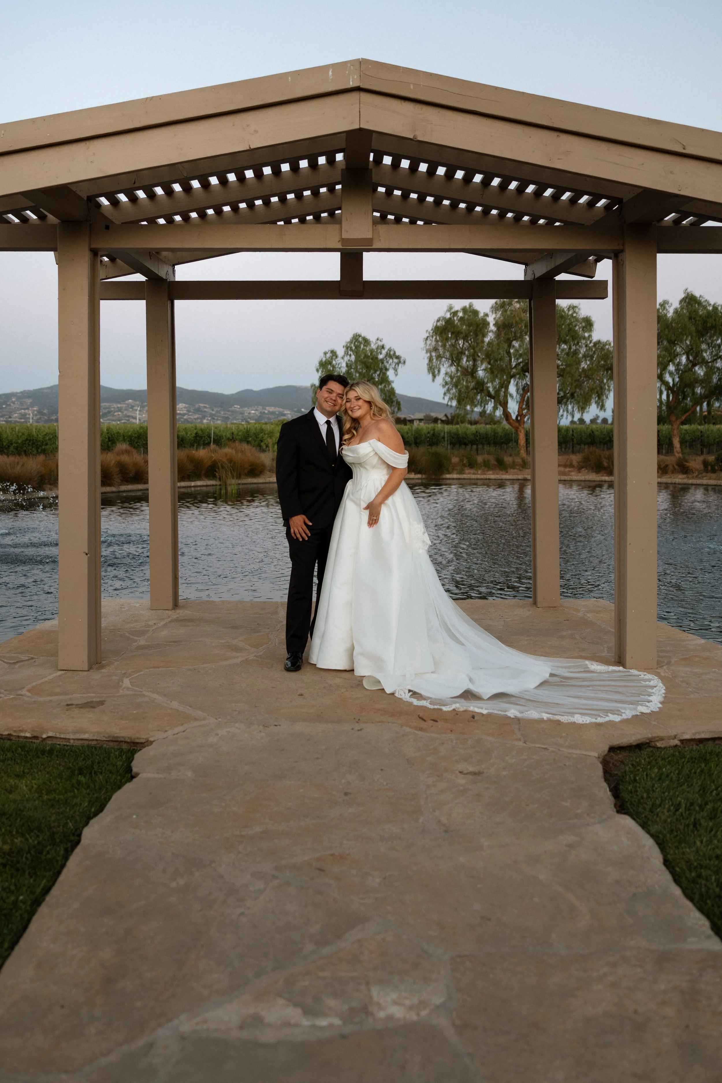 Bride and groom embrace beneath a pergola beside the water with vineyard views