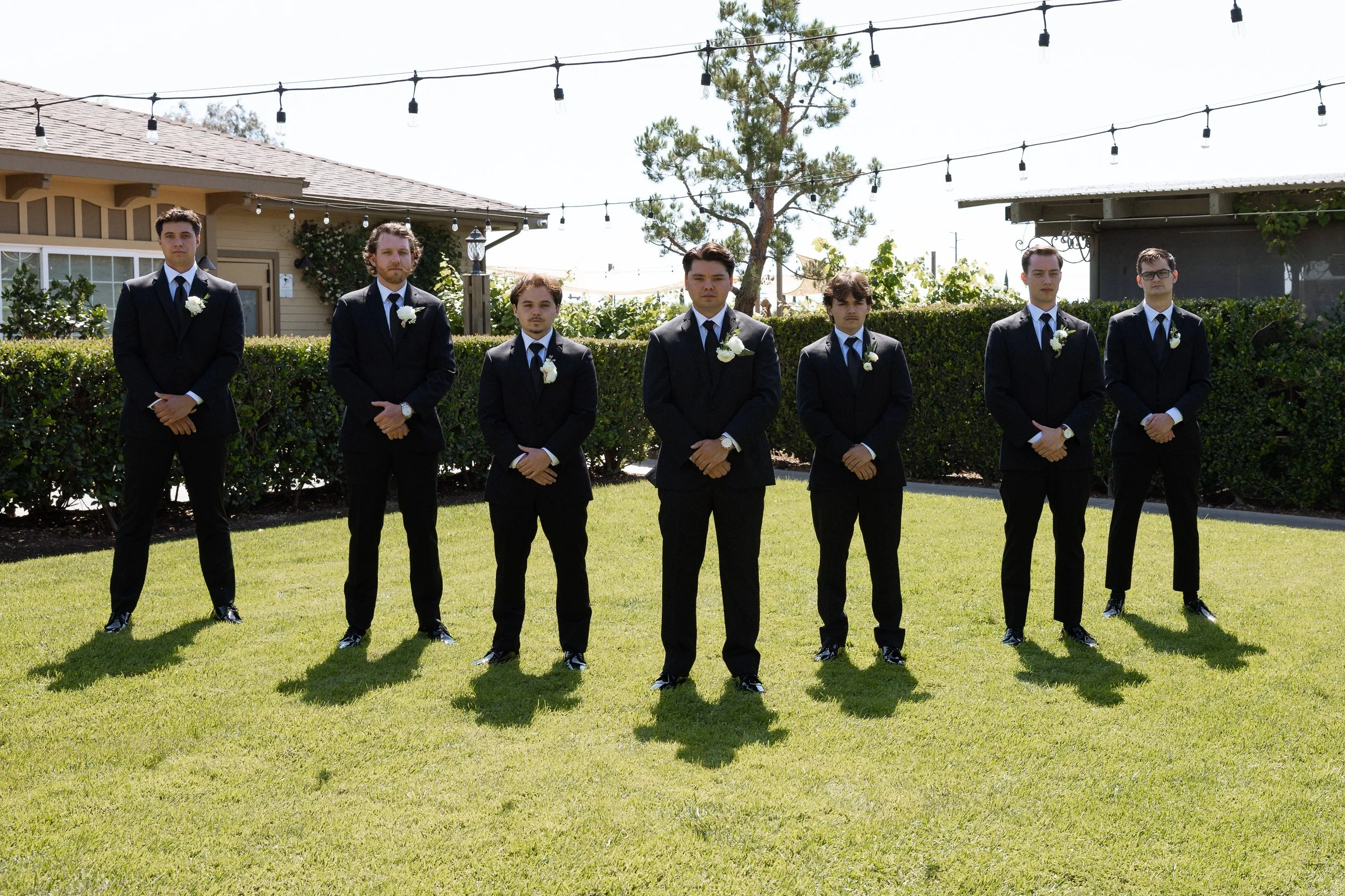 Groomsmen in black suits stand in a row on the lawn before the ceremony
