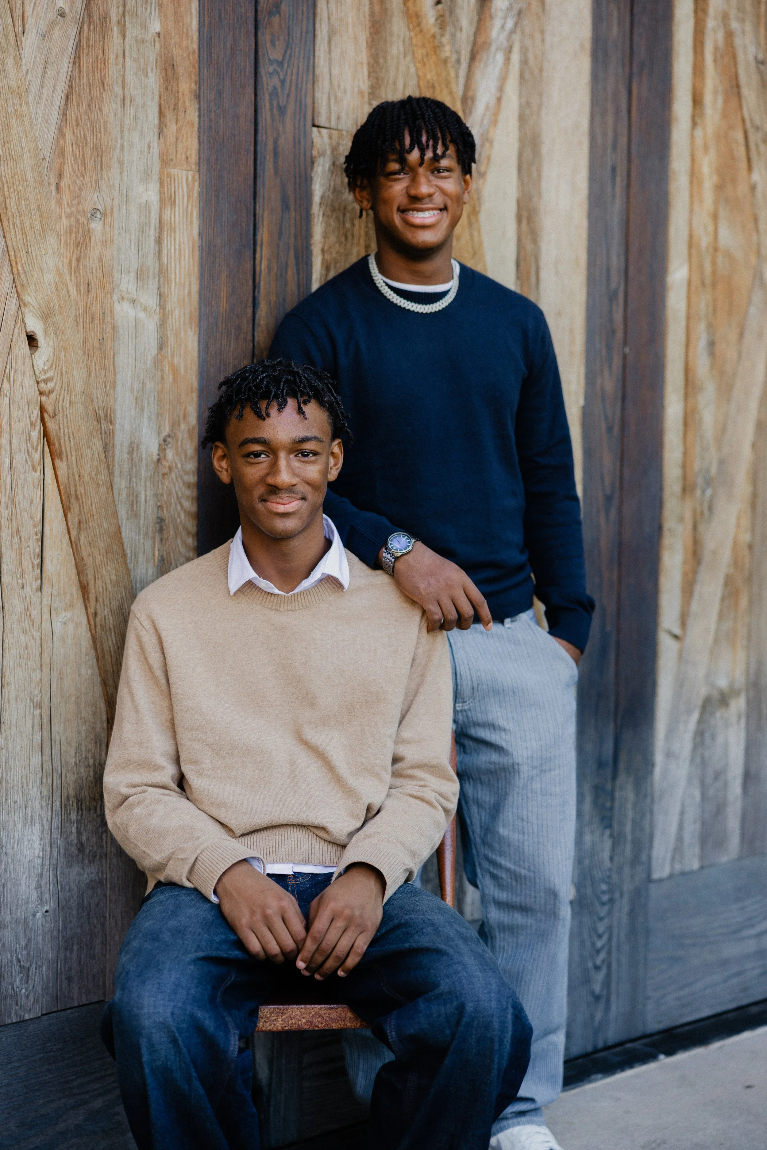Portrait of two brothers, one seated and one standing, posing against a rustic wooden wall at the Great Park.