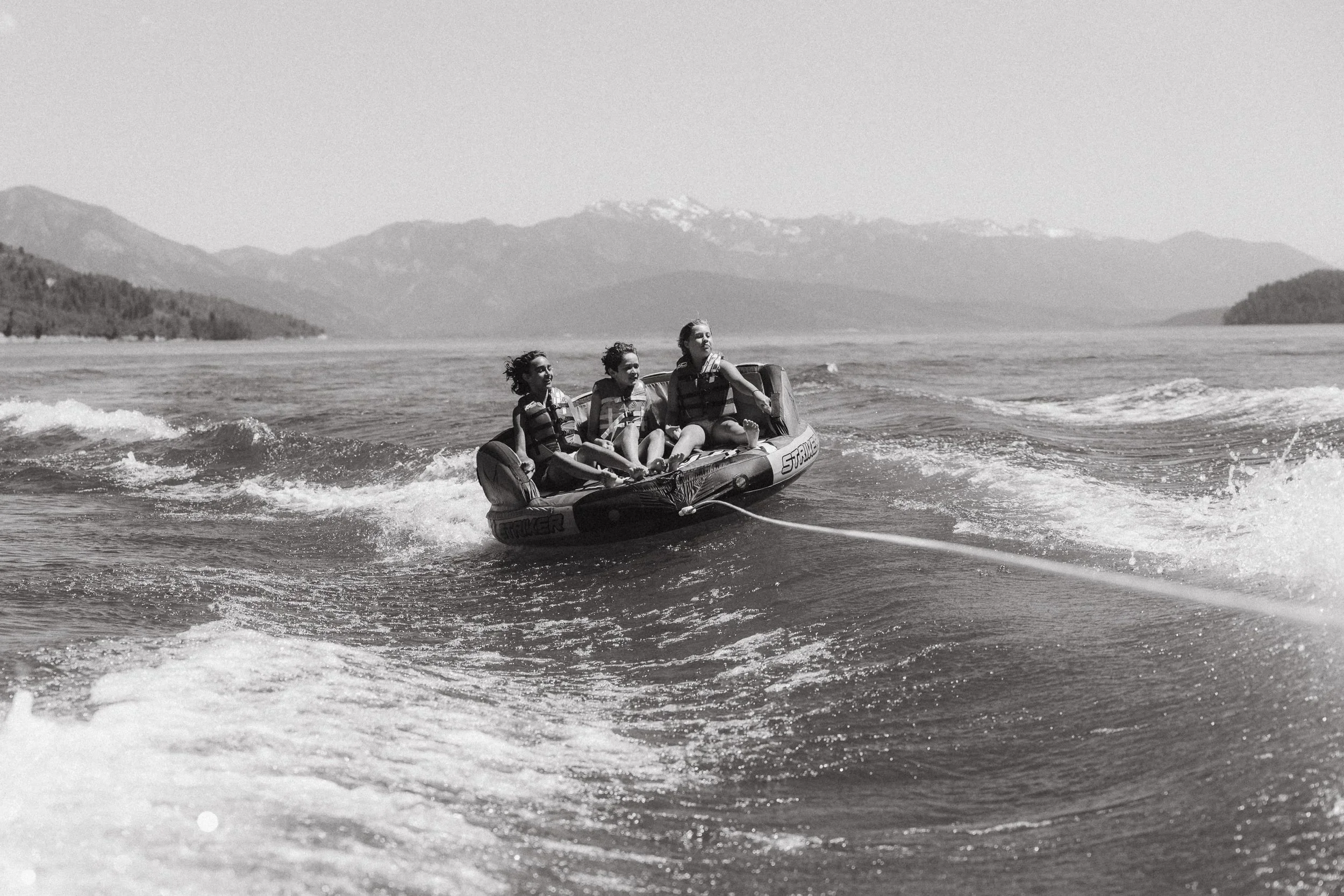Three children laughing on an inflatable tube being pulled by a boat across Palisades Reservoir with the Wyoming mountains in the background.