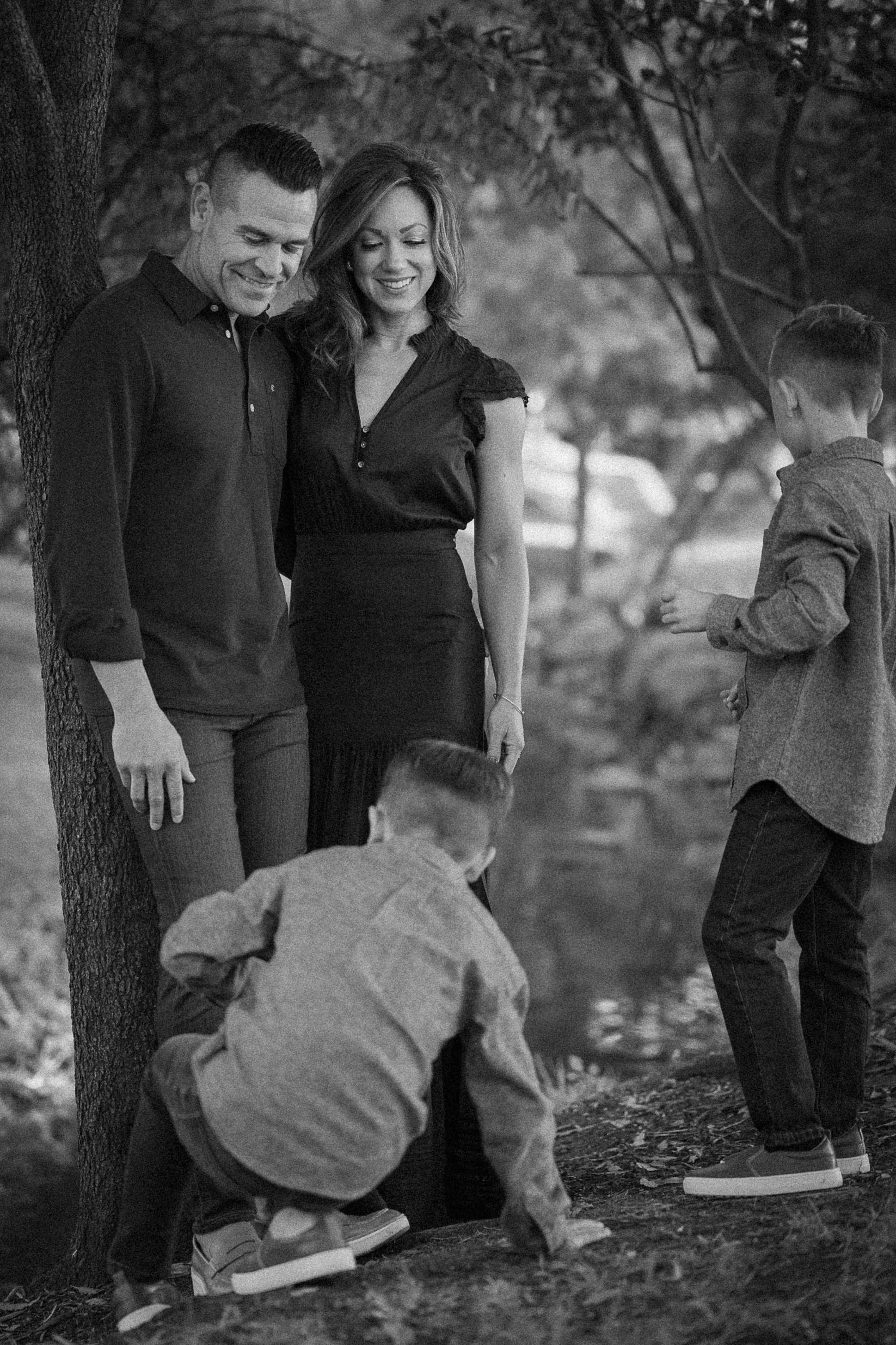 Parents smiling together under the trees while their kids move around nearby.