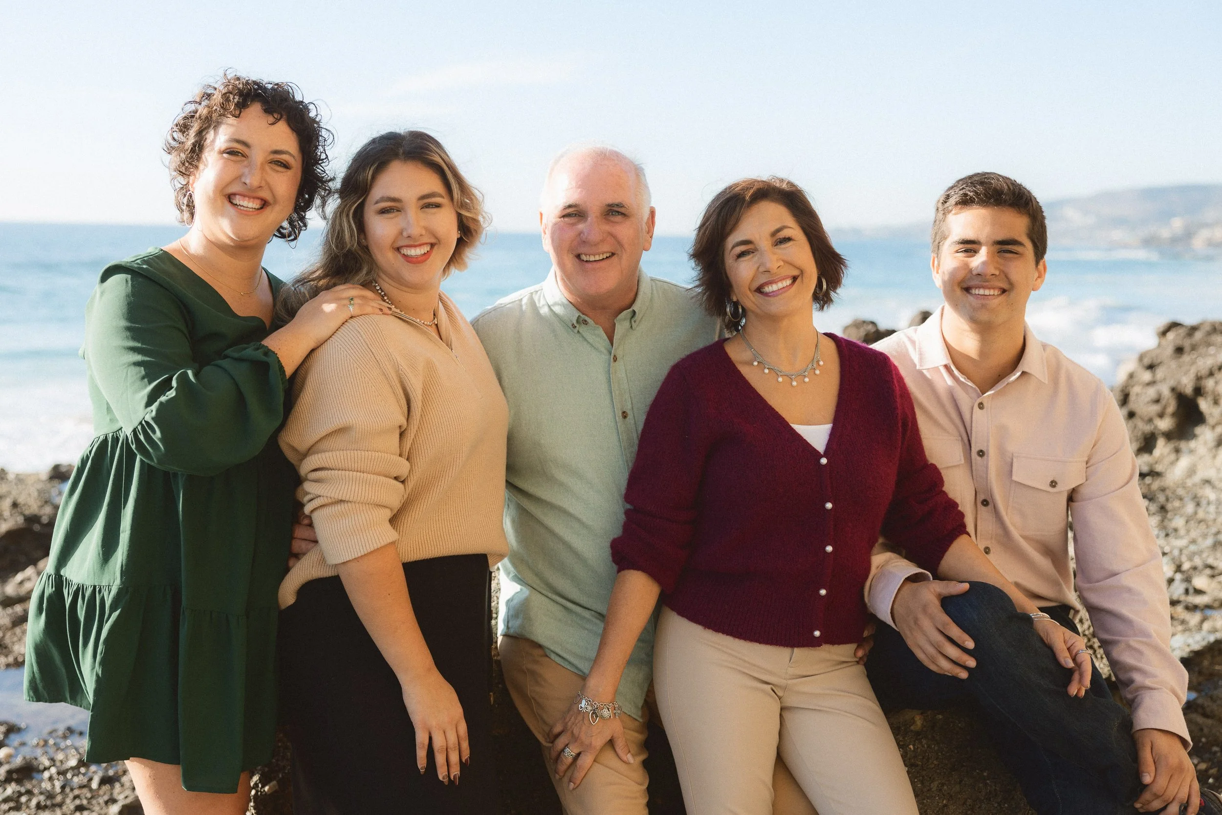 An extended family with adult children smiling for a group portrait on the coastal rocks at Treasure Island Beach in Laguna.