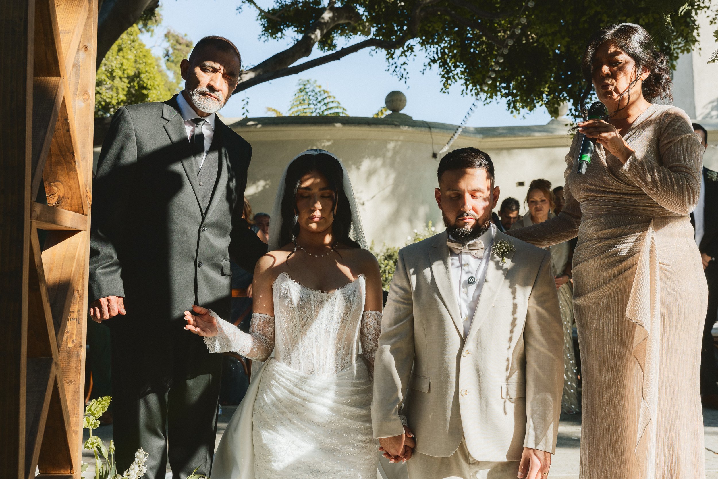 Bride and groom bowing heads in prayer with officiant during ceremony at Los Angeles River Center and Gardens.