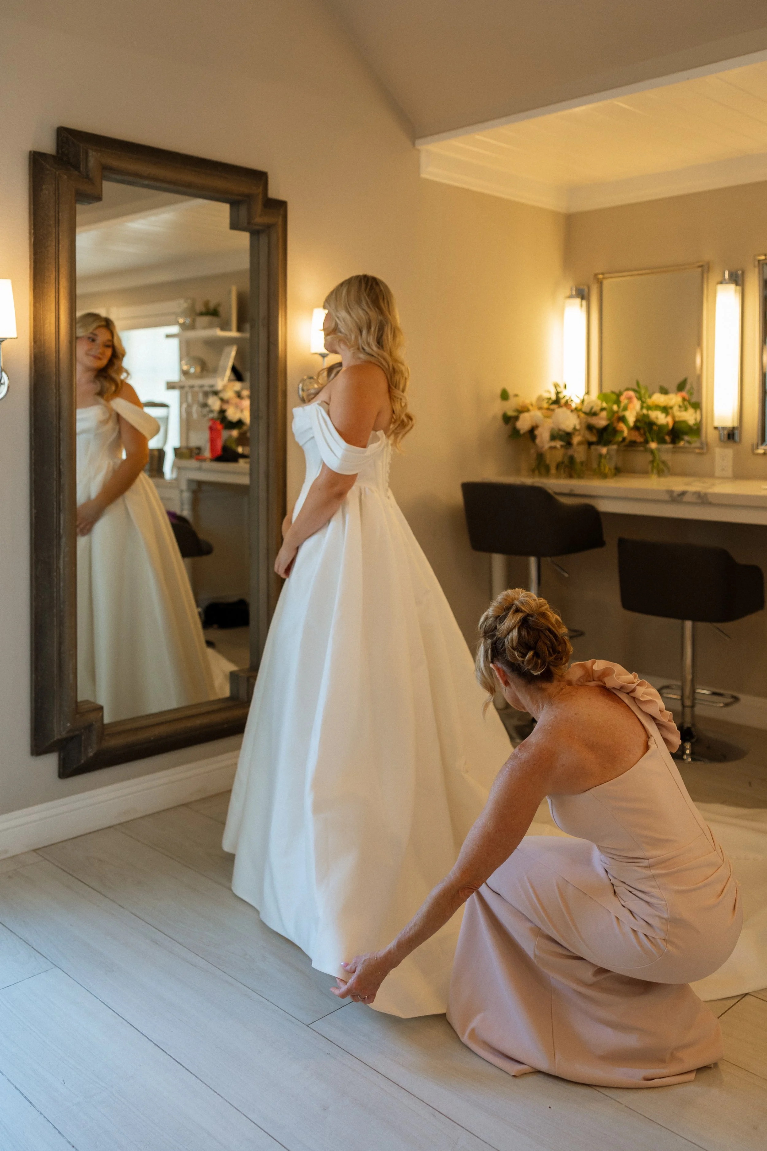 Bride stands at a mirror as a loved one adjusts the train of her gown