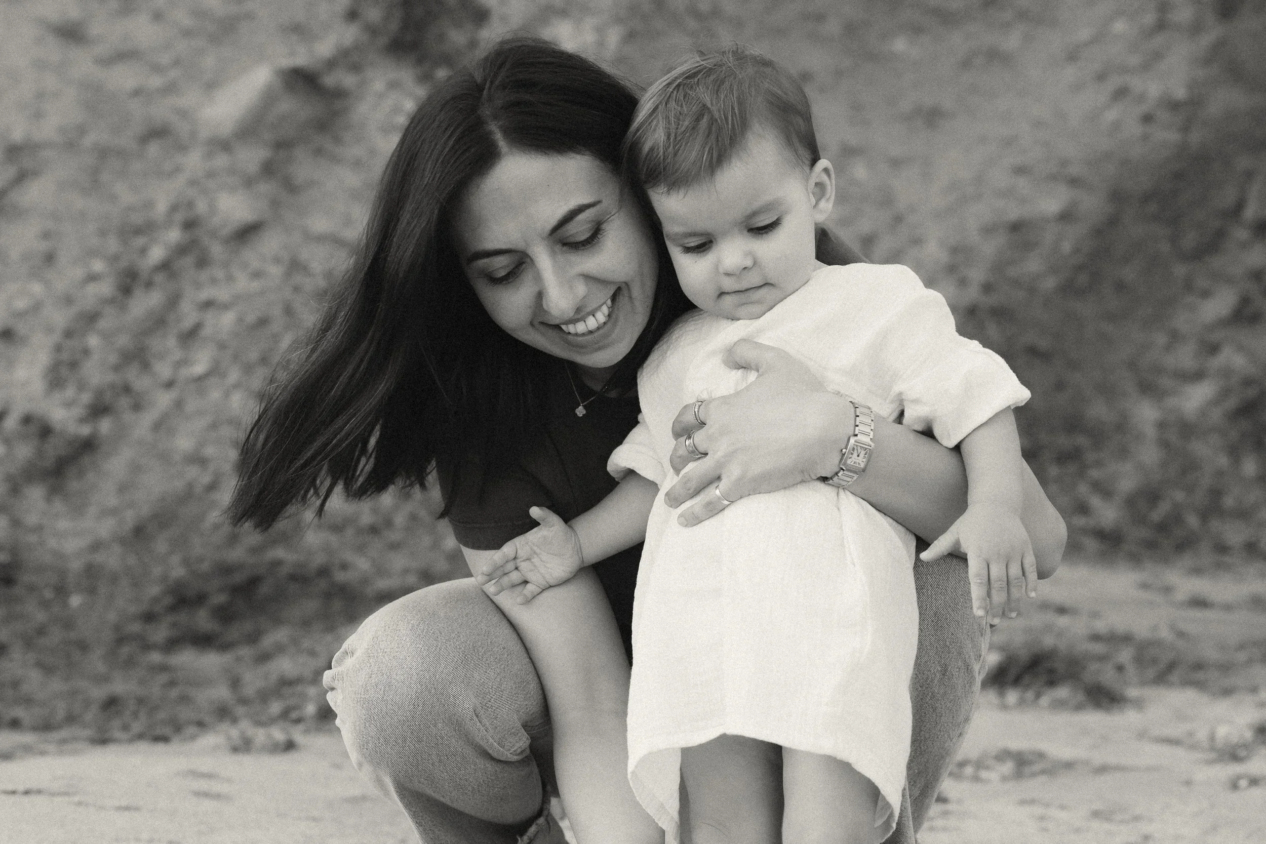 A black and white portrait of a mother holding her toddler daughter while smiling on the beach below the Montage Laguna Beach resort.