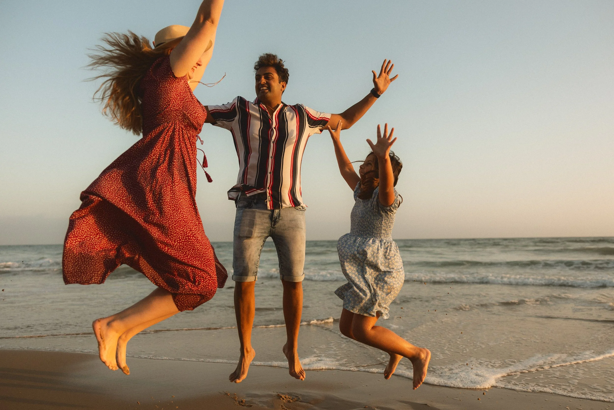A joyful mother, father, and daughter jumping in the air together on the sand during a sunset family photography session at Crystal Cove State Park.