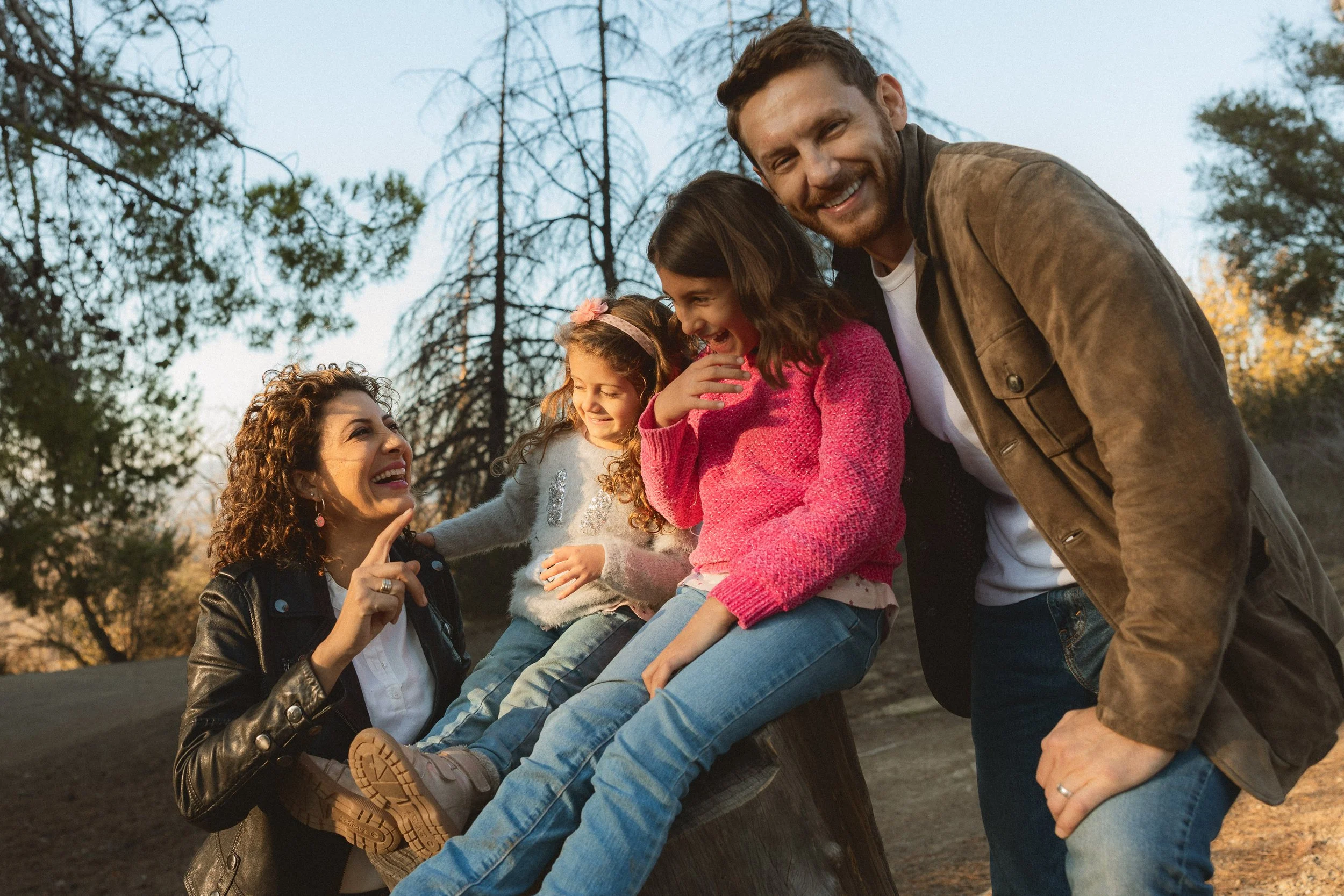 A relaxed family of four sitting on a log and smiling during a sunset session at Debs Regional Park in Los Angeles.