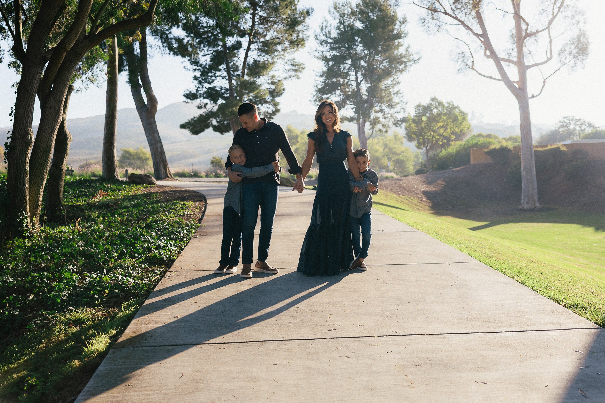 A family of four holding hands and walking away from the camera on a tree-lined path at Turtle Rock Community Park in Irvine.
