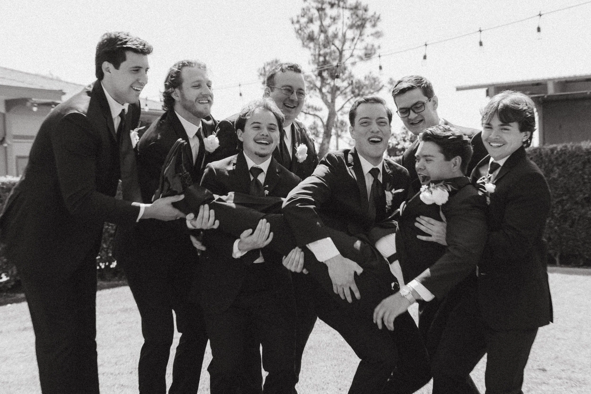 Groomsmen hoist and carry the laughing groom in black and white