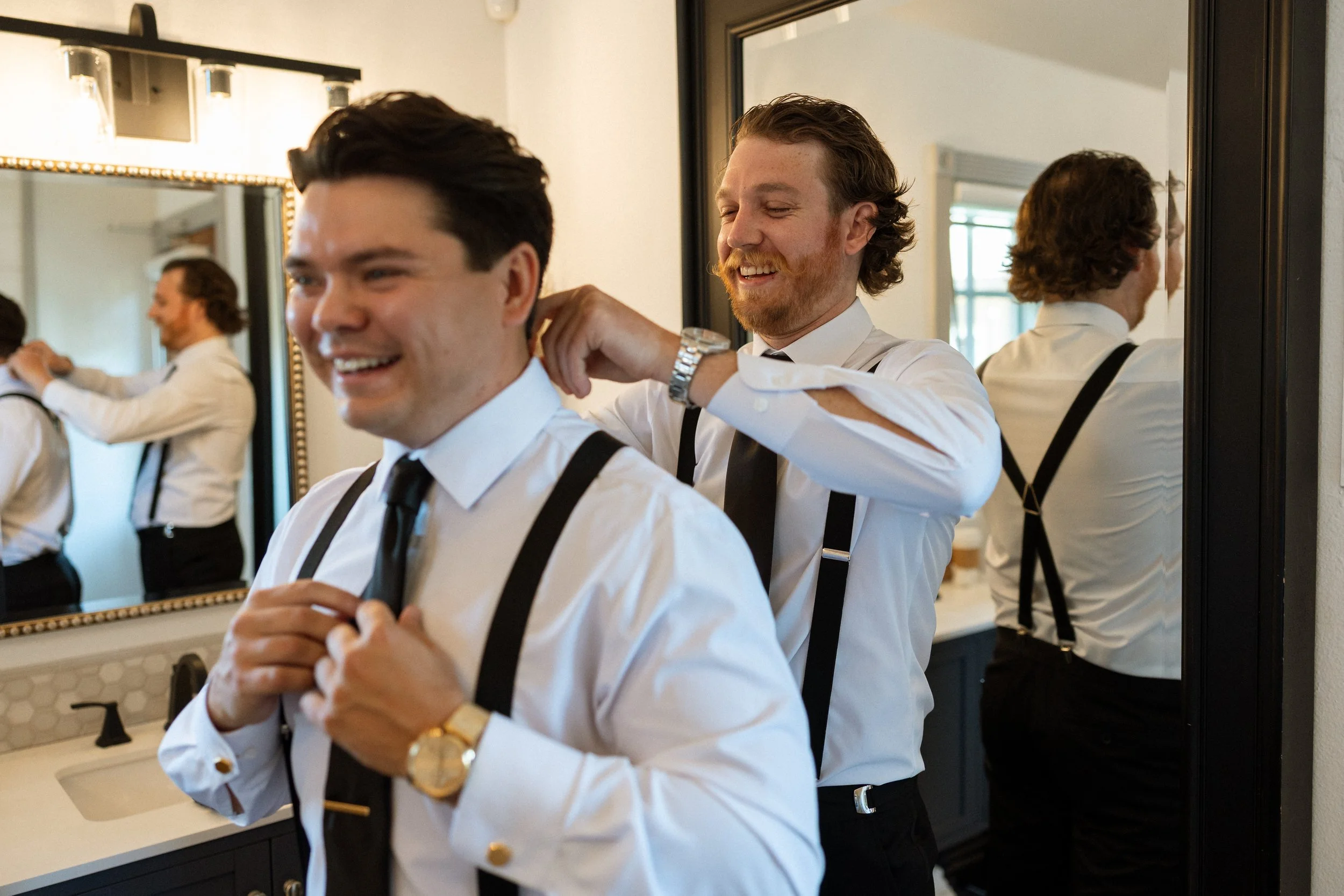 A groomsman helps the groom with his collar as both laugh in the mirror