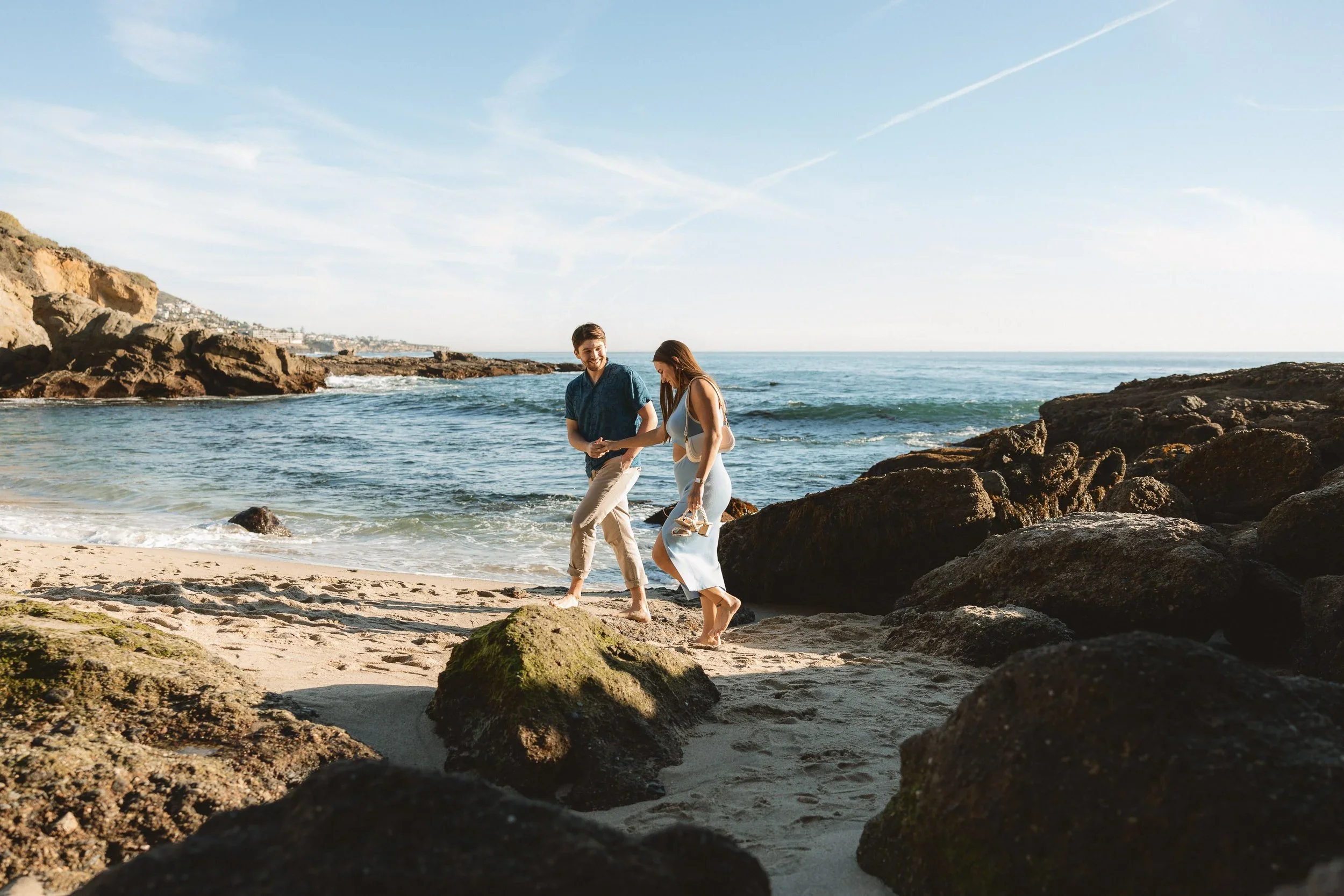 An engaged couple holding hands and walking barefoot on the sand with the rocky cliffs of Montage Laguna Beach in the background.