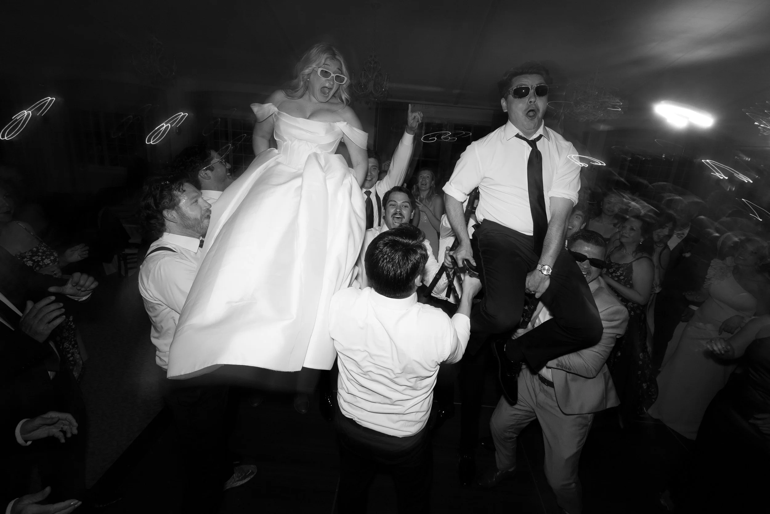 Bride and groom hoisted on chairs above a celebrating crowd in black and white