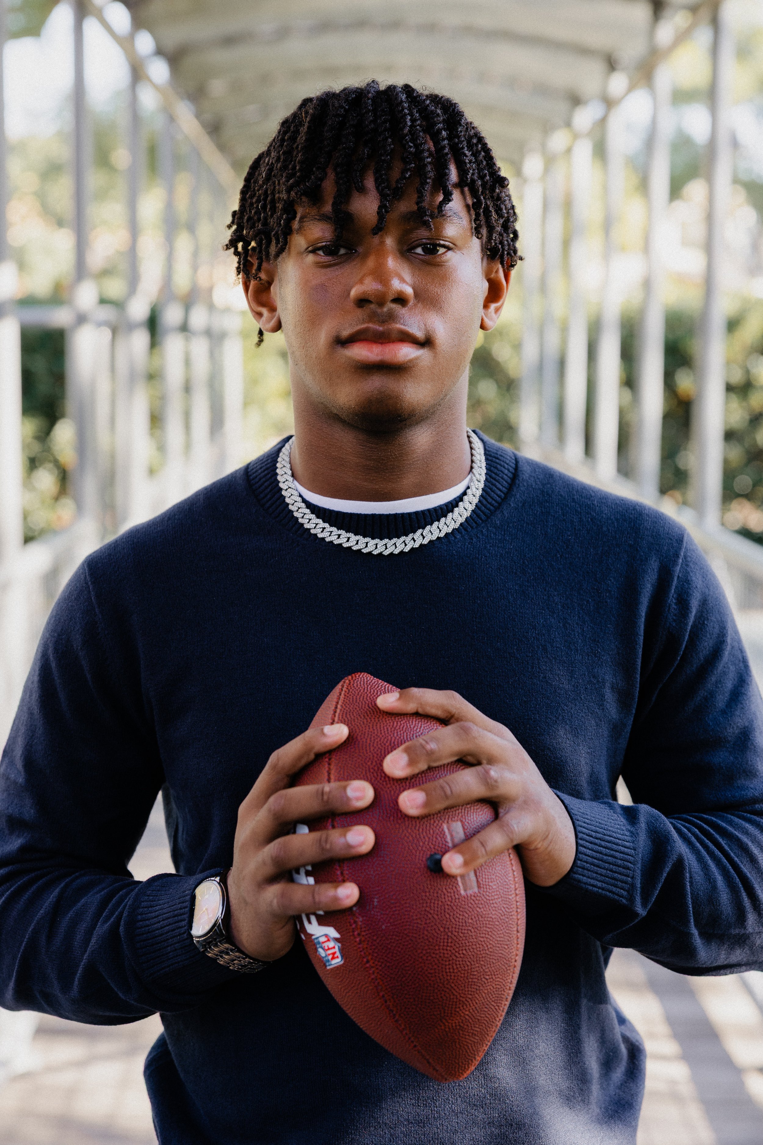 Teenage boy wearing a navy sweater and silver chain holding a football during a portrait session at Great Park Irvine.